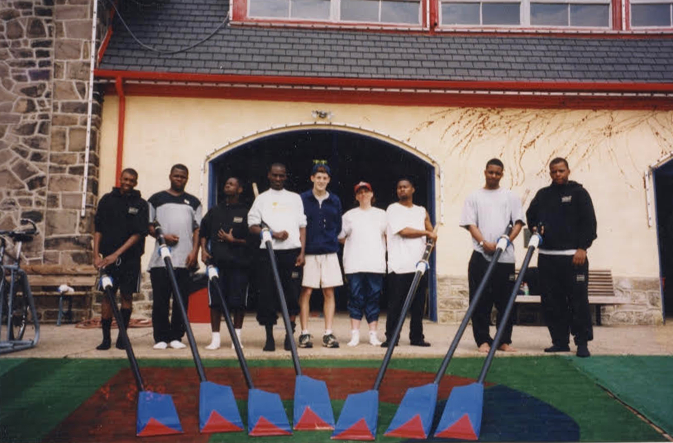 group photo of young black men in front of a boathouse holding oars with their white coach in the middle