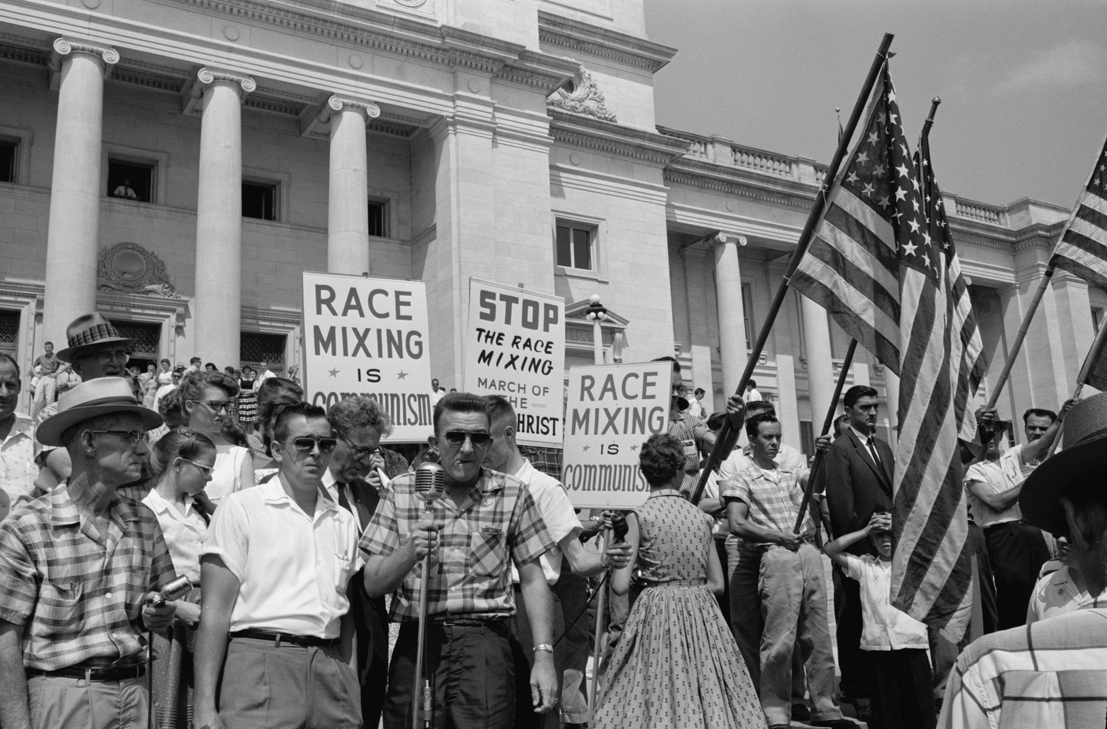 Black and white photo of White people holding signs decrying "race mixing" and American flags at a protest on the steps of a government building