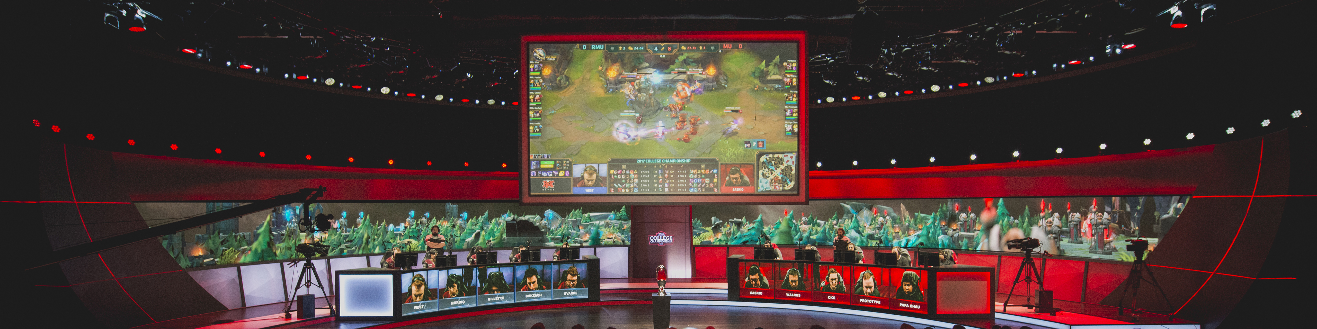 a wide shot of an esports arena featuring a large television screen hanging over a stage with one team of video game players on the left and another team on the right