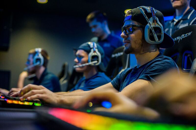 an e sports competitor from columbia college wearing glasses, a backwards hat, and a headset with microphone sits with his hands on a keyboard in front of a computer in a row of other gamers