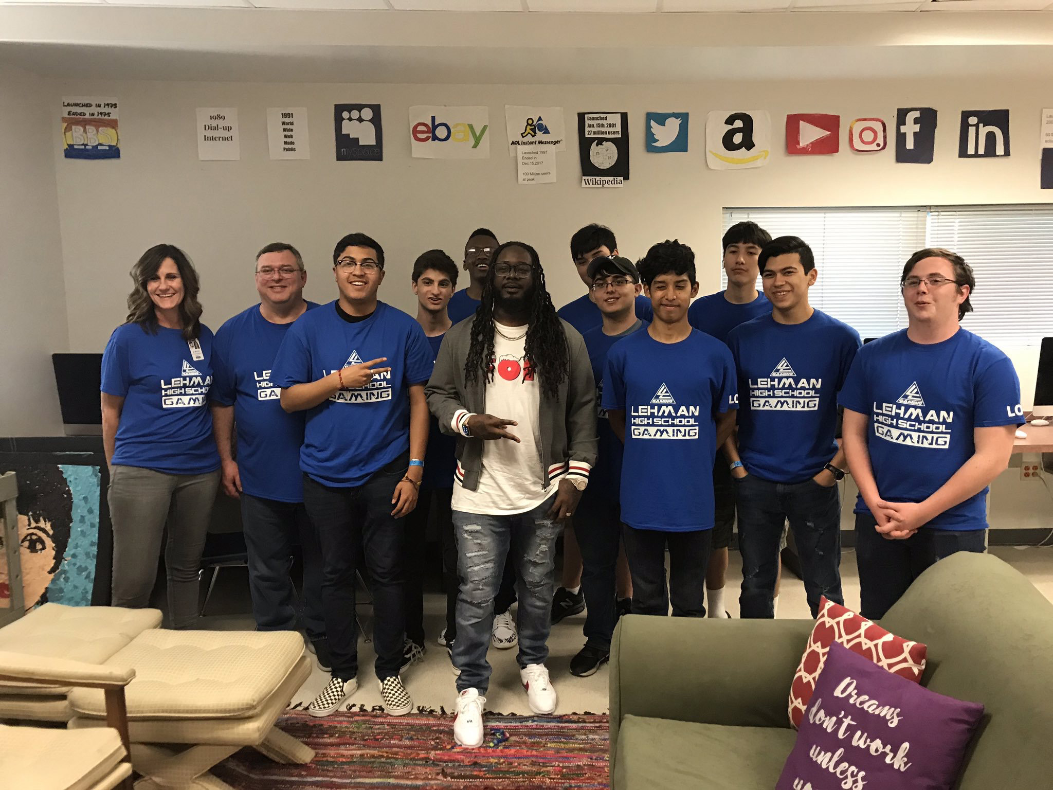 males students and male and female teachers from the lehman high school gaming club pose for a photo in a room with hand drawn tech company logos posted on the wall behind them