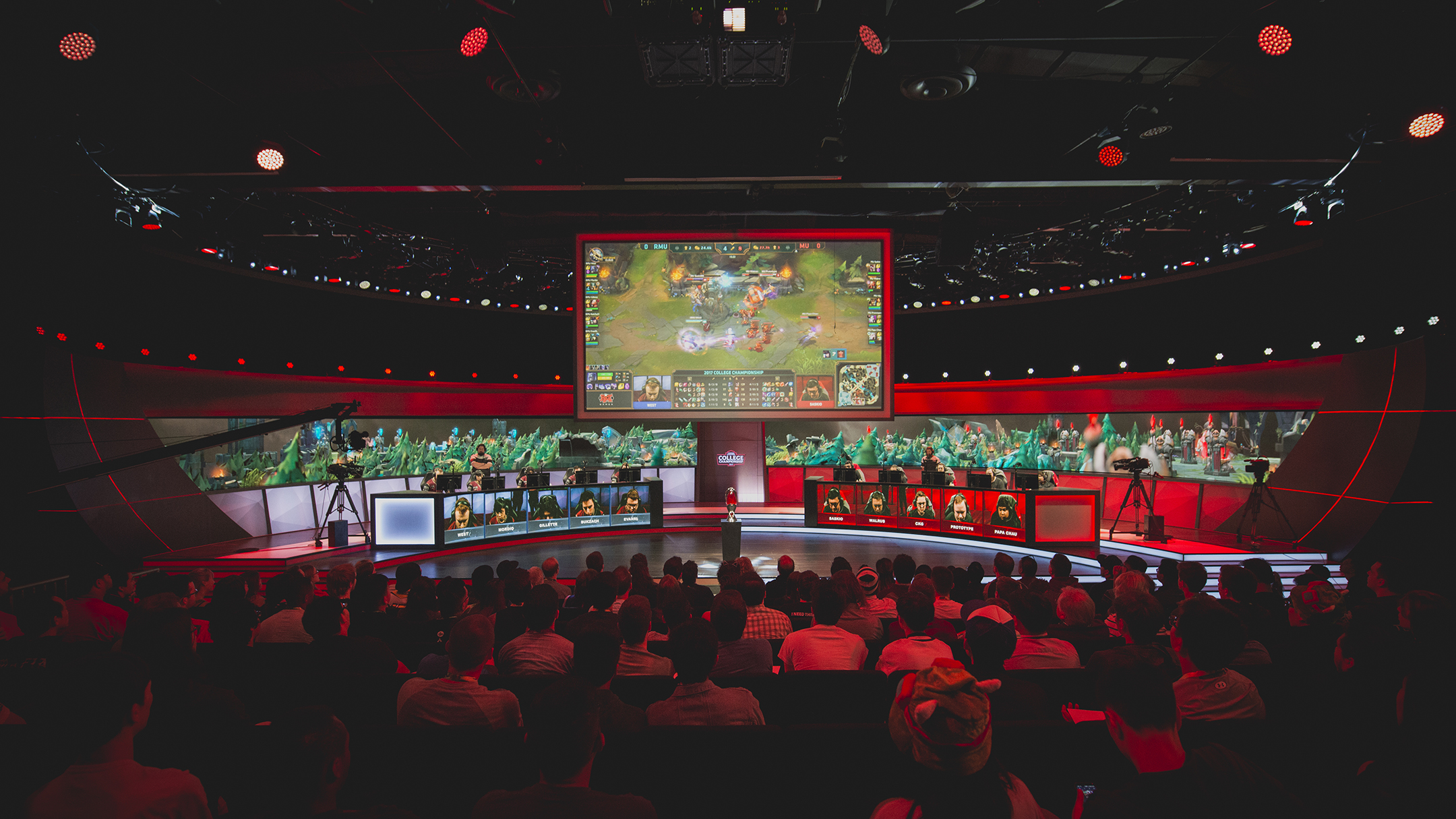 a wide shot of an esports arena featuring a large television screen hanging over a stage with one team of video game players on the left and another team on the right