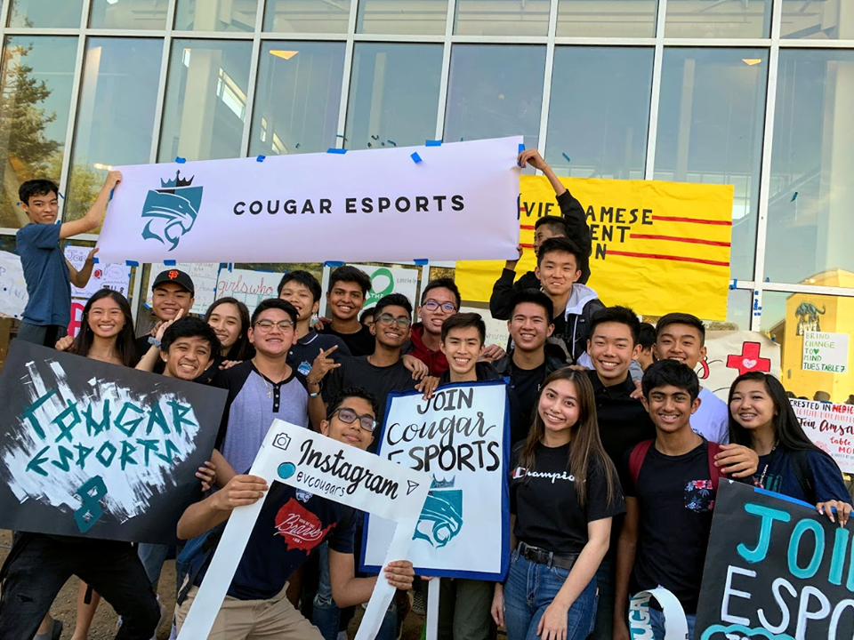 A group of students who are part of their school's e sports team gather outside a building holding signs and banners promoting playing e sports