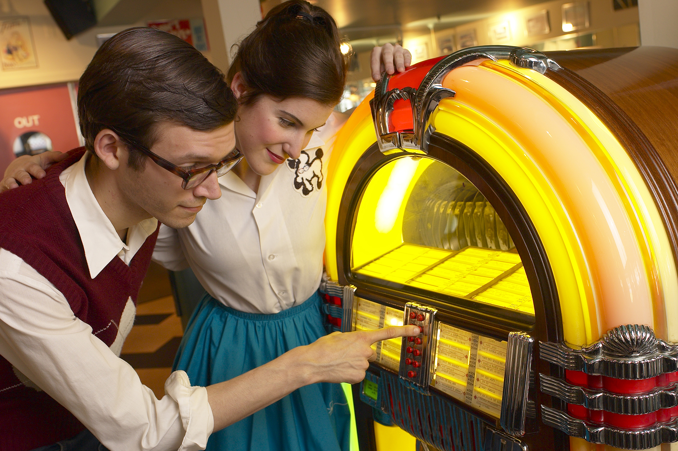 a couple dressed like they're from the 1950s stand in front of a jukebox while the young man presses a button