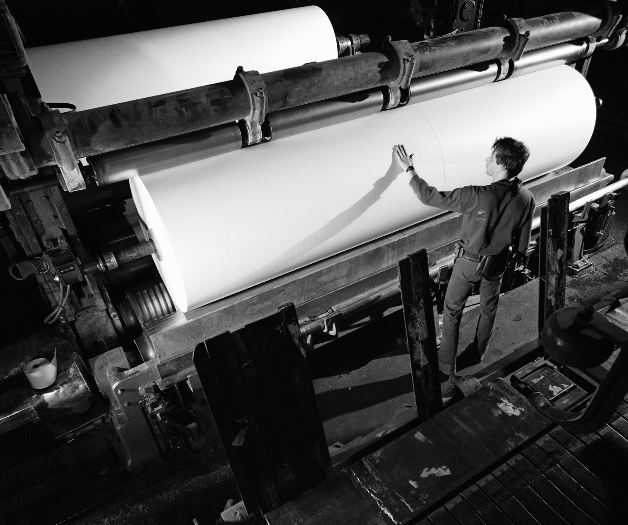 a black and white photo of a male worker looking at and touch a roll of paper in a paper mill