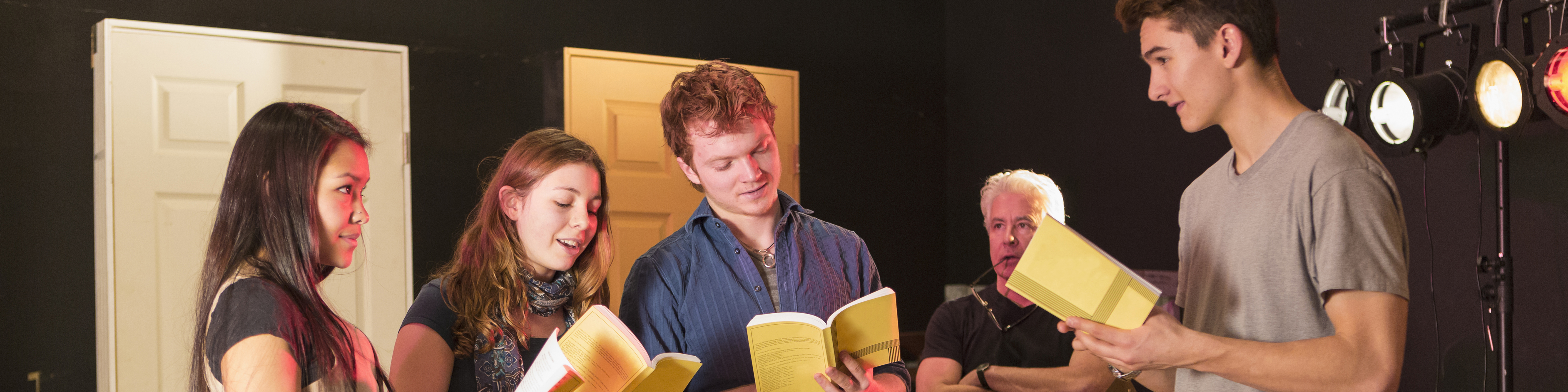 a group of male and female high school students stand on a stage reading from scripts as they rehearse for a play