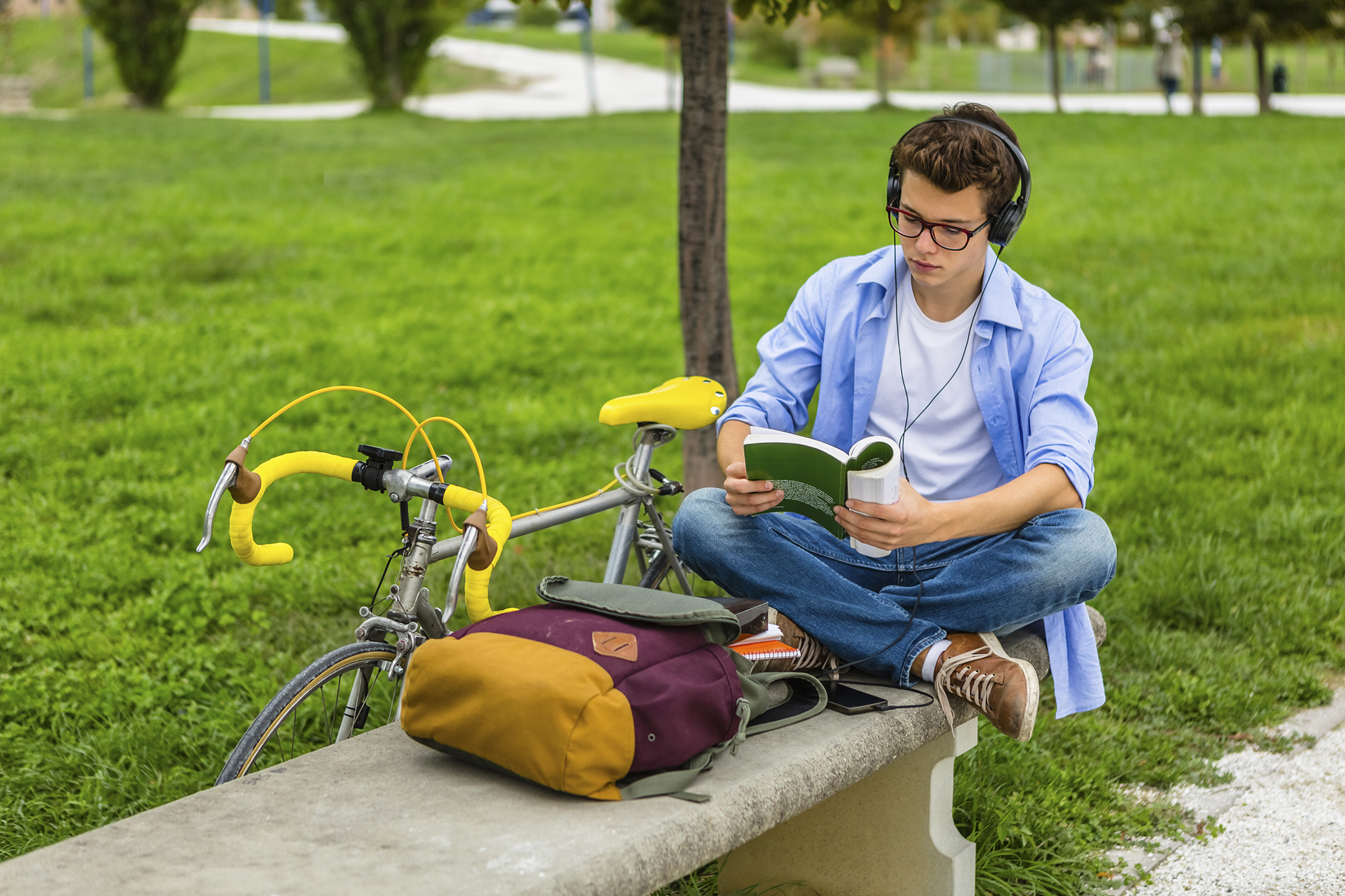 a college student sitting on a bench reading a book while wearing headphones and guarding his bike