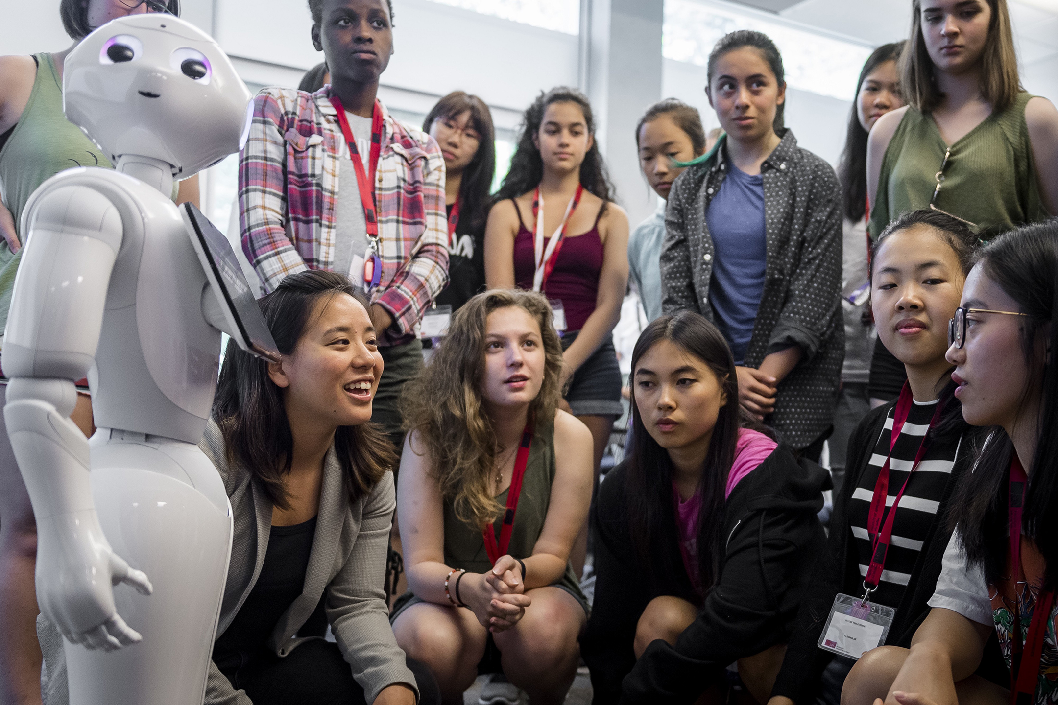 A group of a dozen female high school students listen to an adult woman talk about an human-looking artificial-intelligence-based robot
