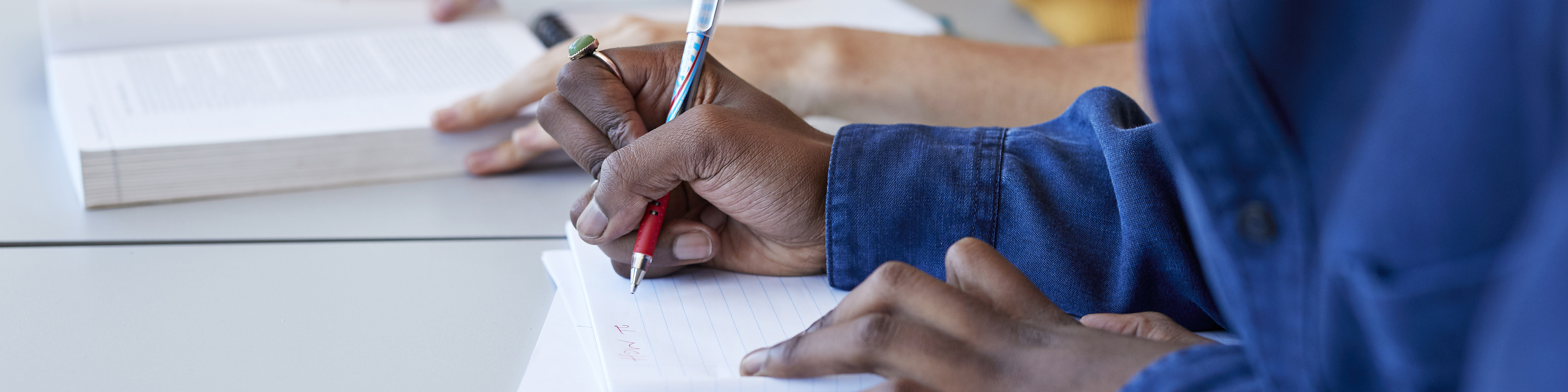 a close up of a pair of hands on a desk, with the right hand clutching a red pen that is being used to write on a blank piece of paper in a notebook