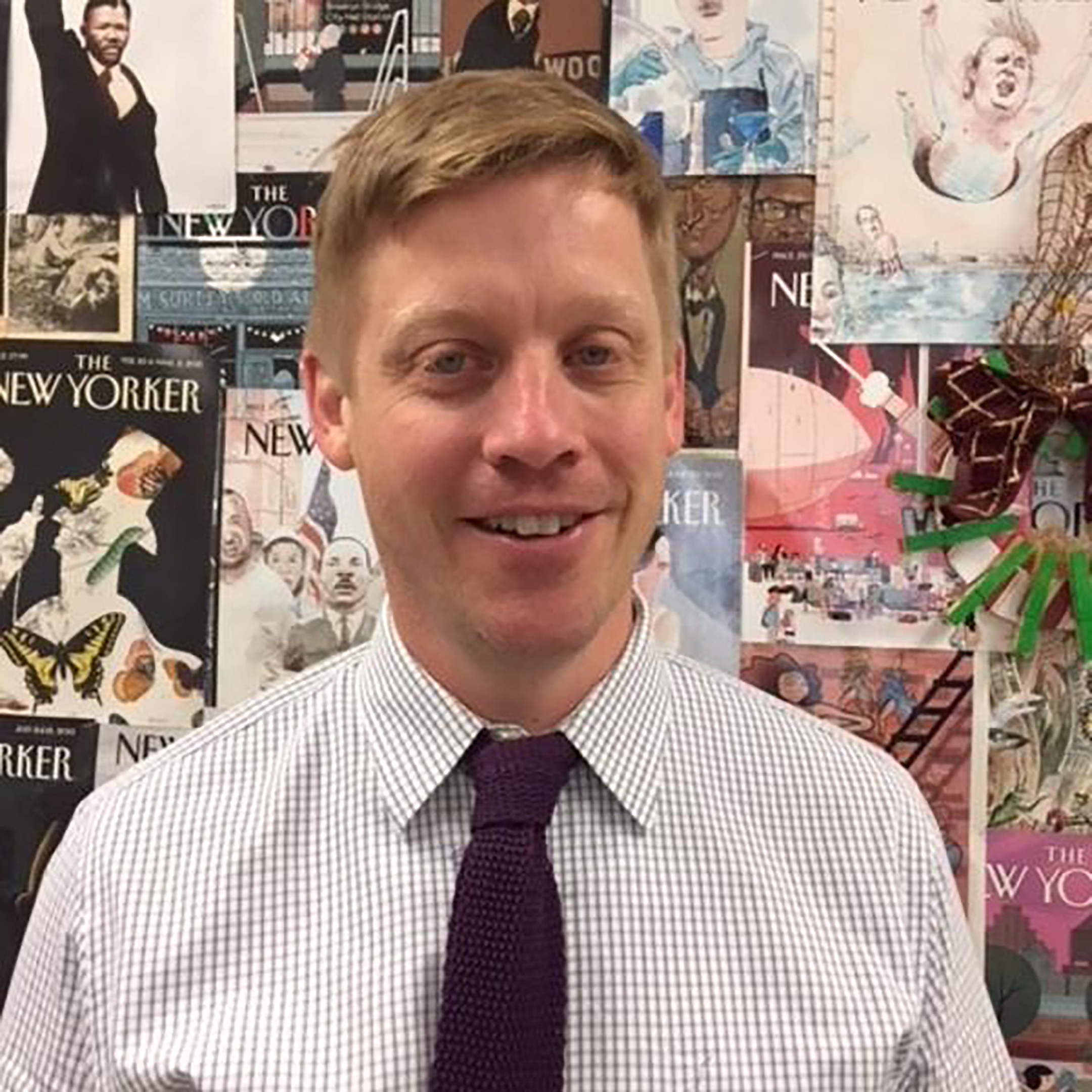 a profile photo of teacher brendan widness, taken from the chest up, showing him wearing a white shirt and dark tie and standing in front of a wall papered with covers of the new yorker magazine