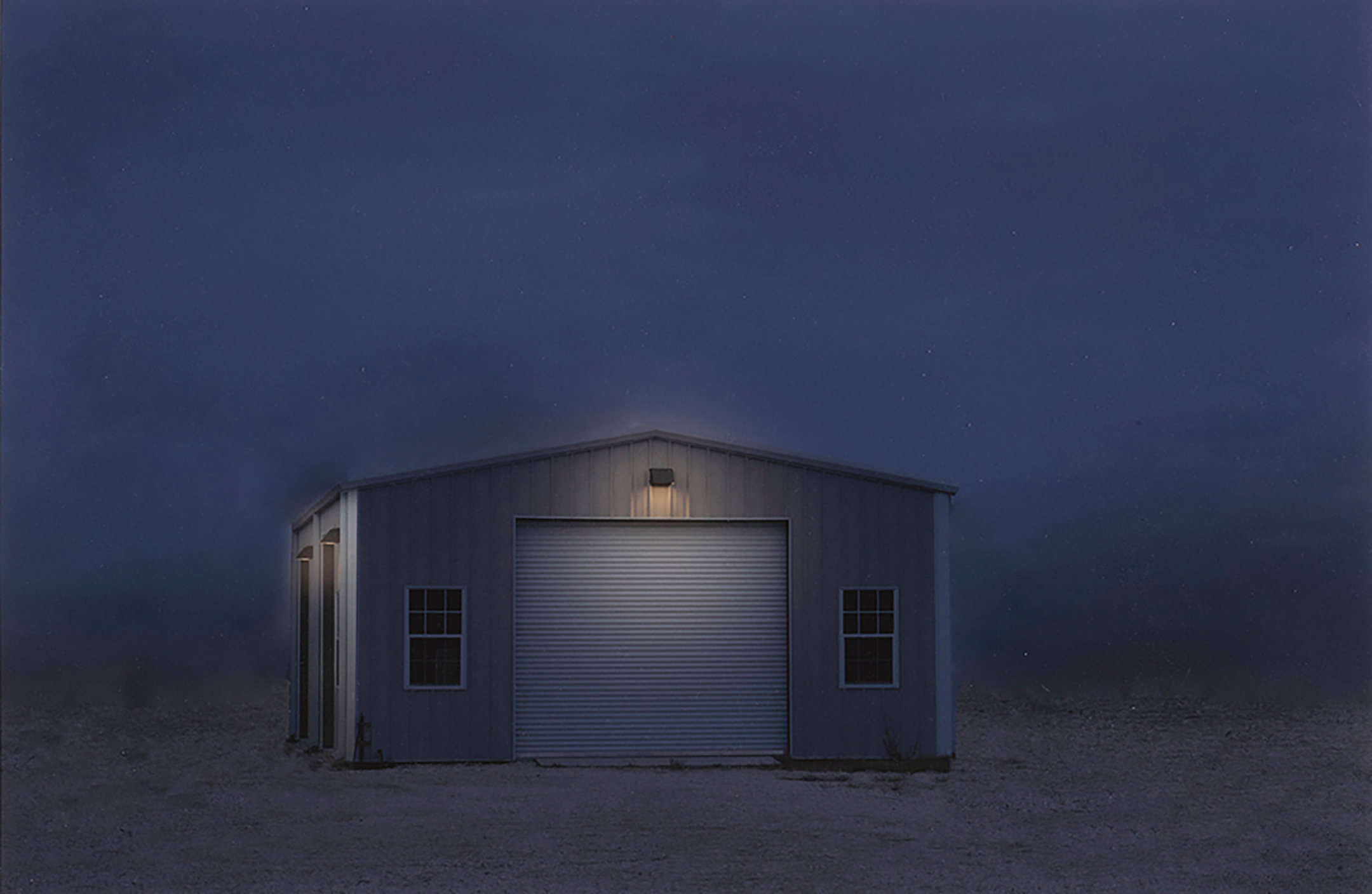 A photograph taken at twilight of a solitary single-story beige shed with a slightly pitched roof and a large, closed garage door and two small windows on either side illuminated by a small lamp hanging above the door 
