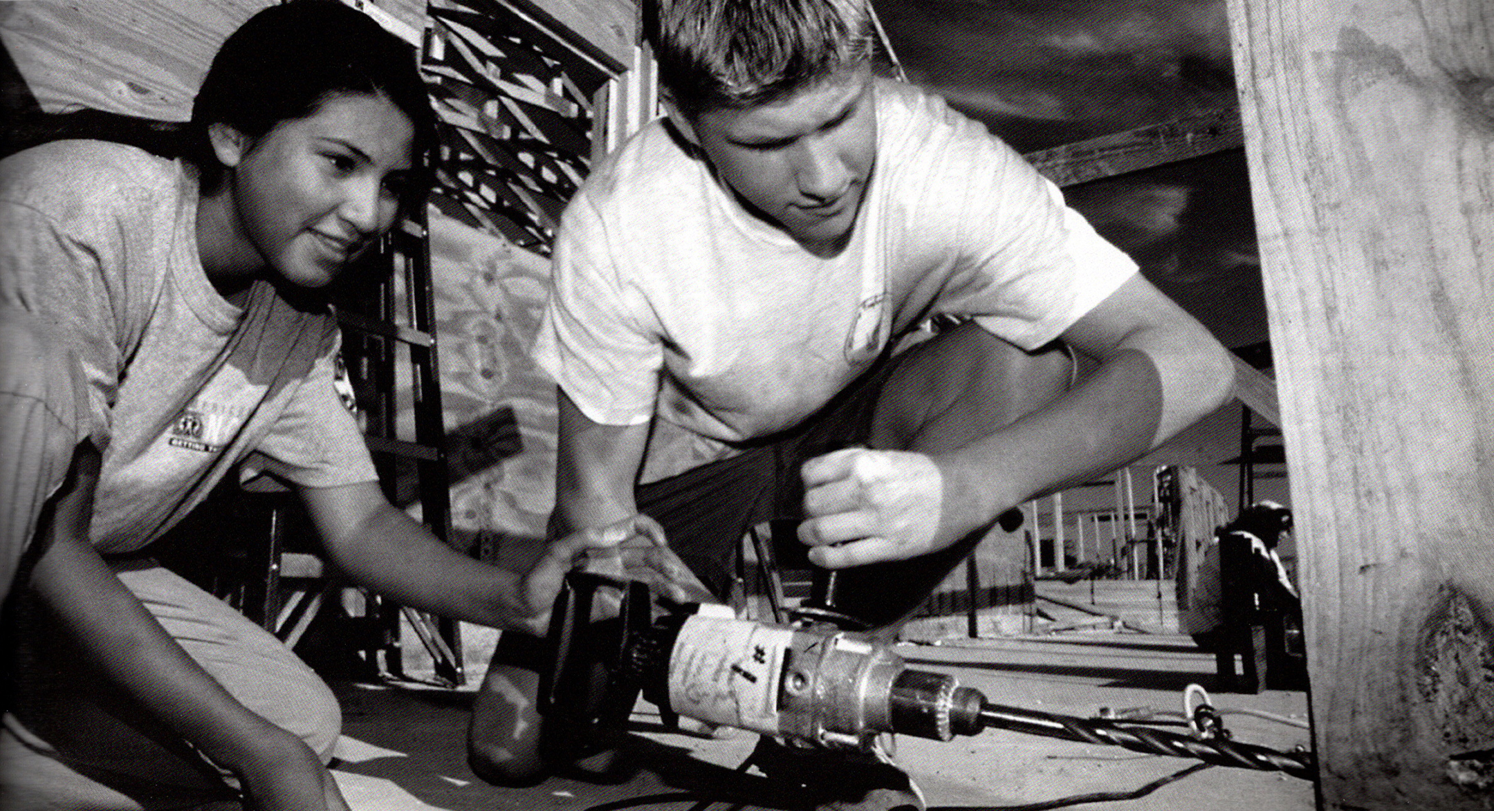 black and white photo of two students, one male and one female, drilling a hole into a beam of wood at a worksite