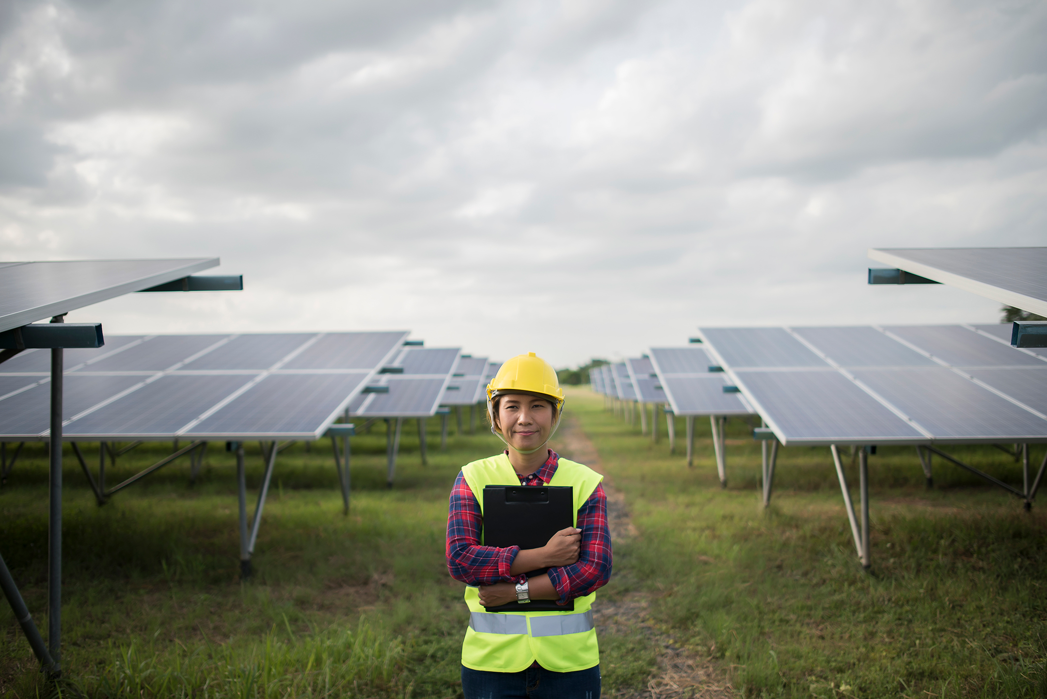 a woman in a hard hat and work vest stands in a field, clutching a cipboard, with two rows of solar panels behind her