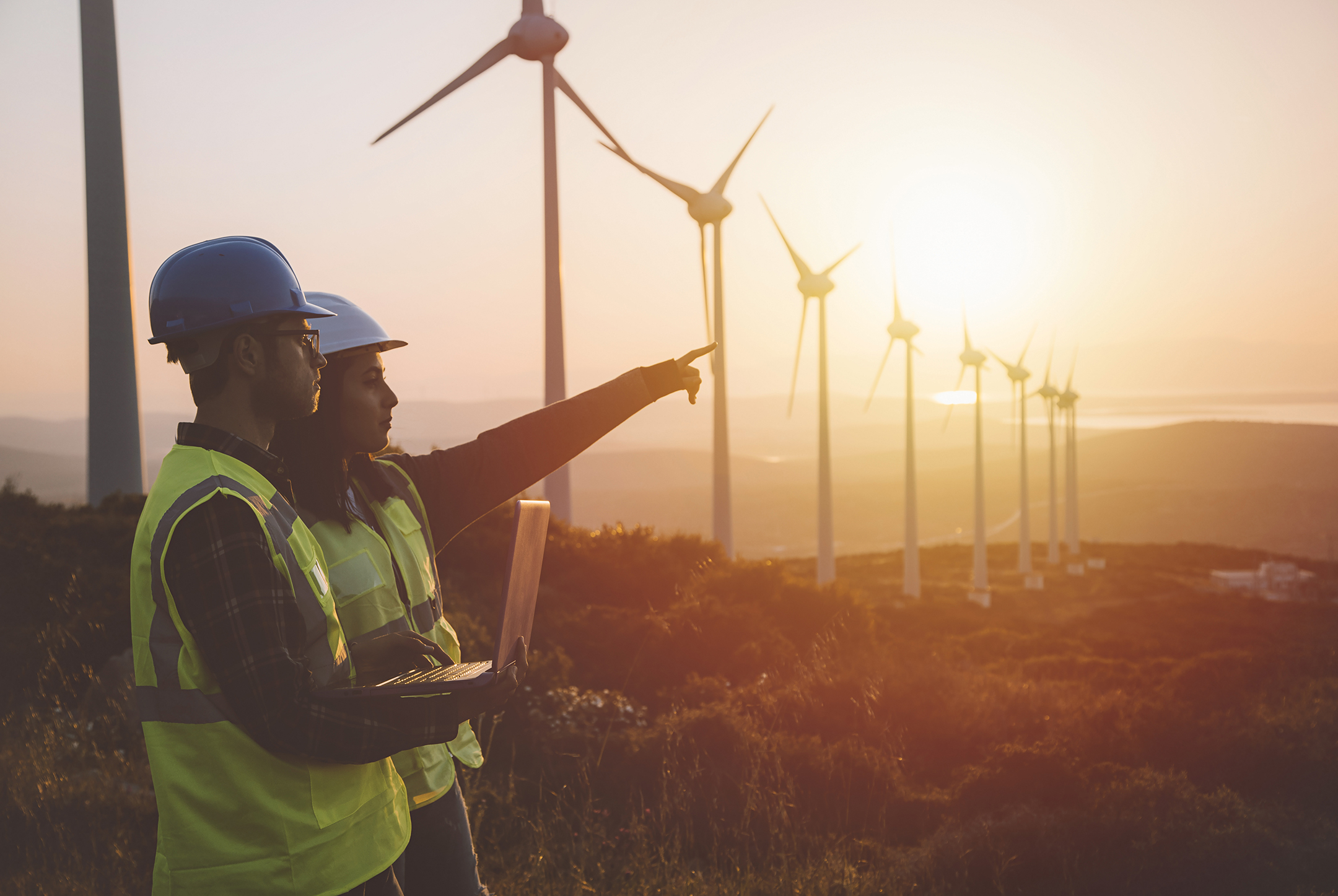 a man and a woman wearing hard hats and work vests stand in front of a row of wind energy turbines in a field during sunset, the woman pointing to the right 