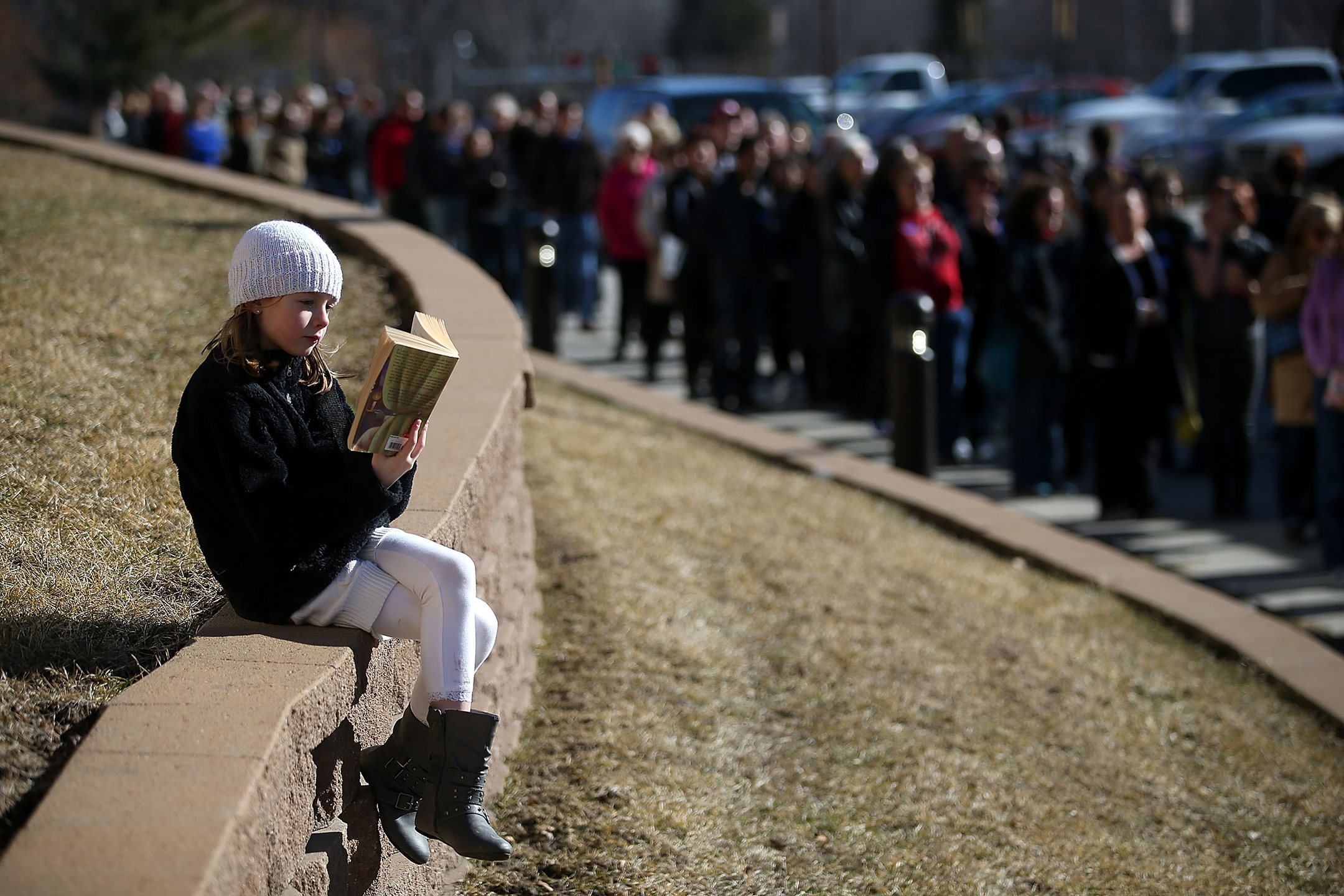 a young woman in a coat and hat sits alone on a wall reading while a line of people wait to enter a political rally
