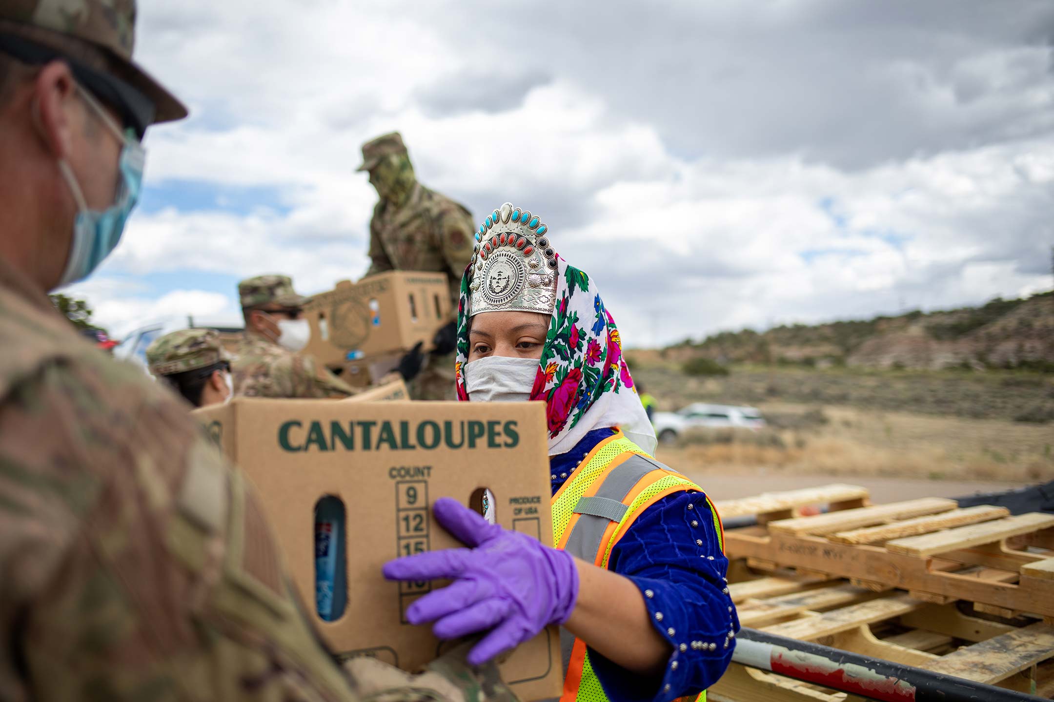 a woman in a traditional navajo headdress and wearing purple gloves and a neon yellow work vest over her clothes accepts a box of food from a soldier.