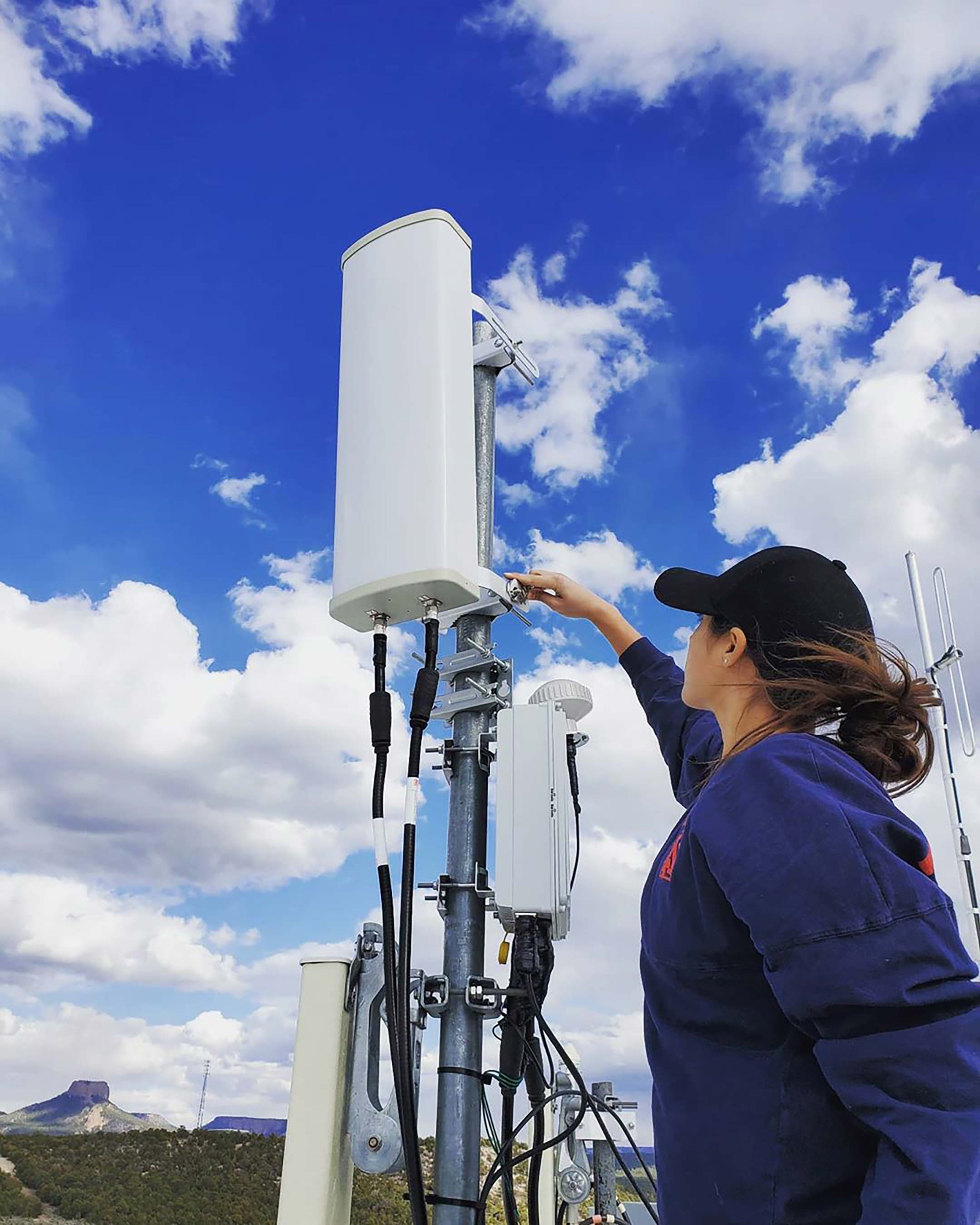 a woman in a blue jacket and cap works on a piece of technology equipment mounted on a pole
