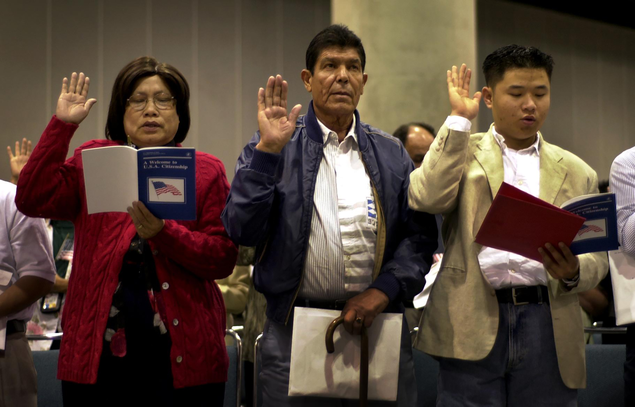 three people, from left, a woman and two men, raise their hands as they take the oath of citizenship