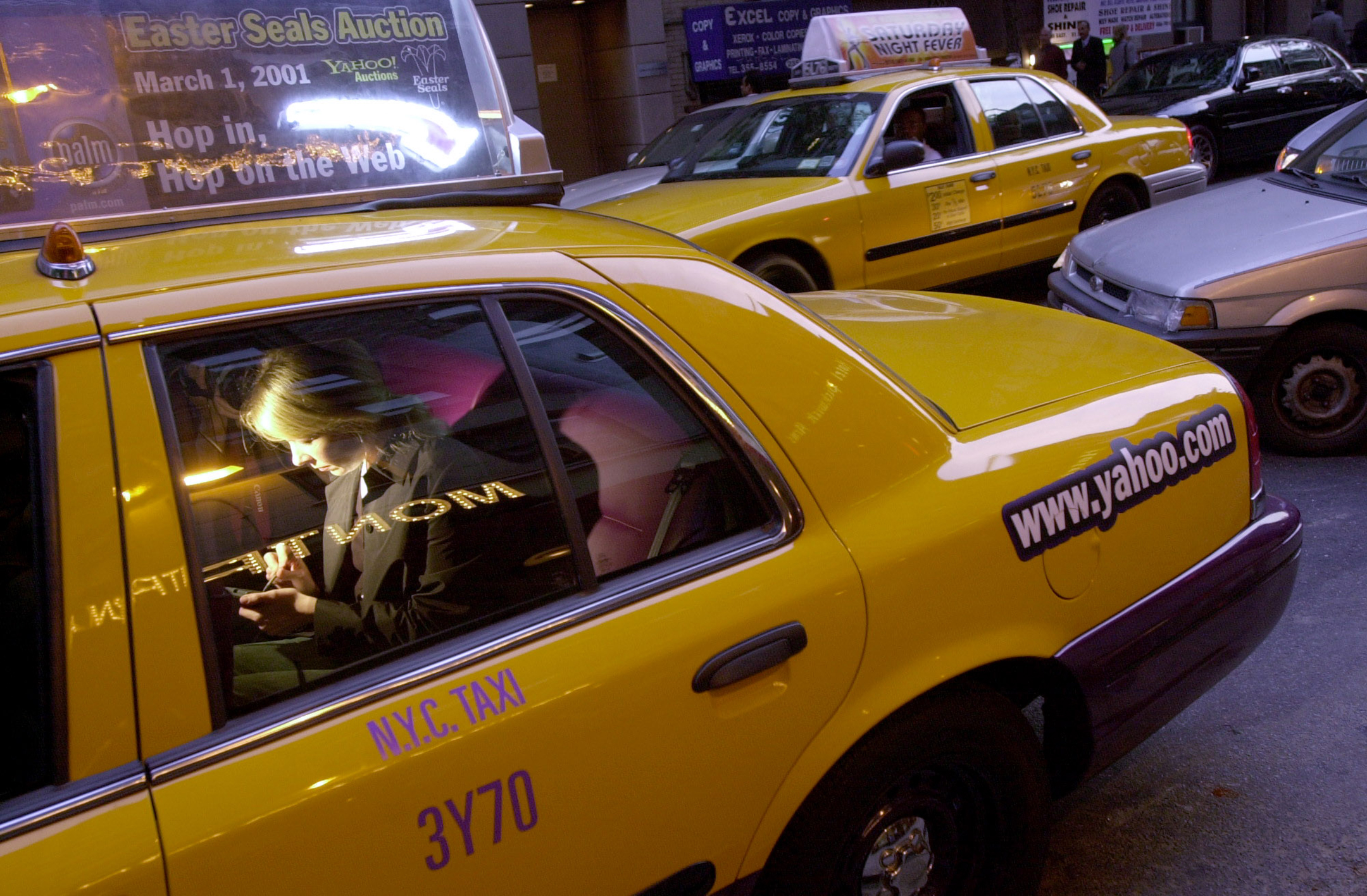 seen from the outside, a woman sitting in the back of a yellow taxicab looks at her device