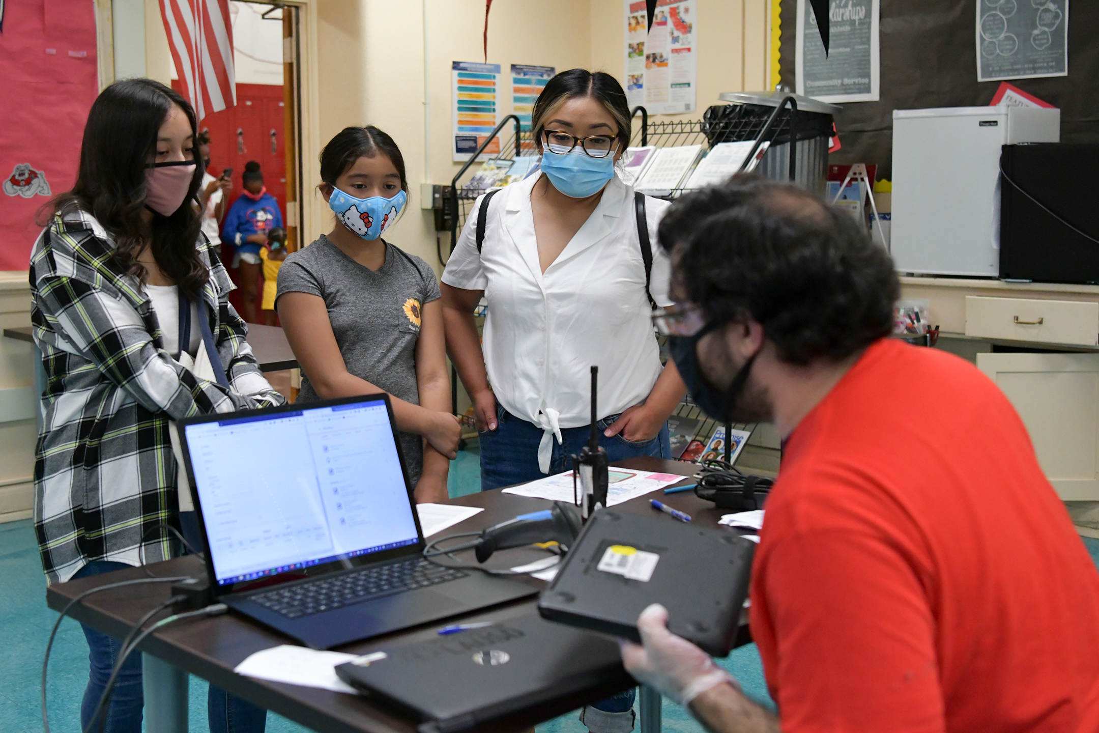 A mother and her two children, all wearing face masks, stand in front of a school administrator who is preparing to loan them a laptop computer