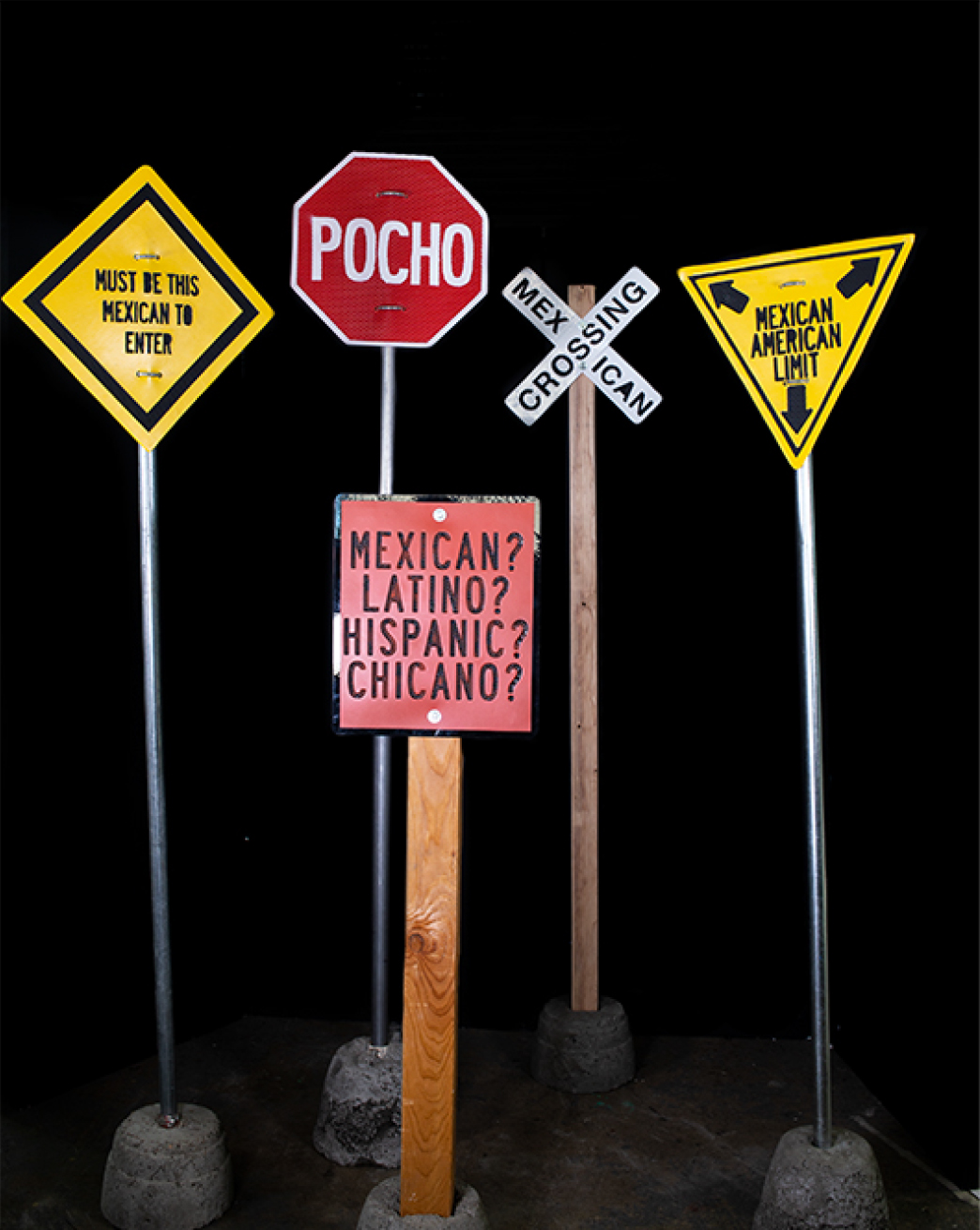 Photo of five fake road signs against a black background with messages aimed at Mexicans and Mexican Americans, like Pocho and Must Be This Mexican to Enter