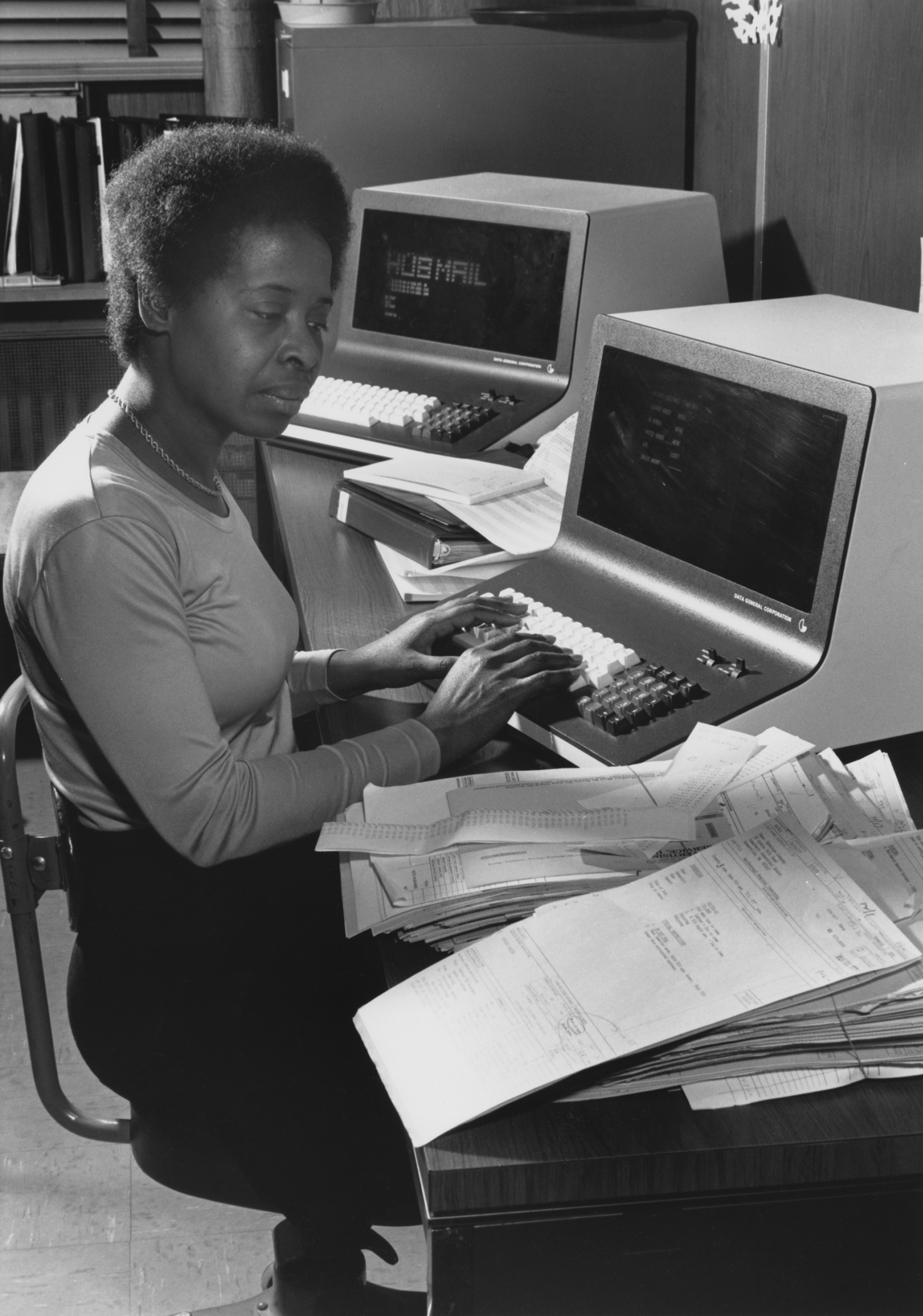 a black and white photo of a woman sitting at a desk with two computer terminals and a stack of paper
