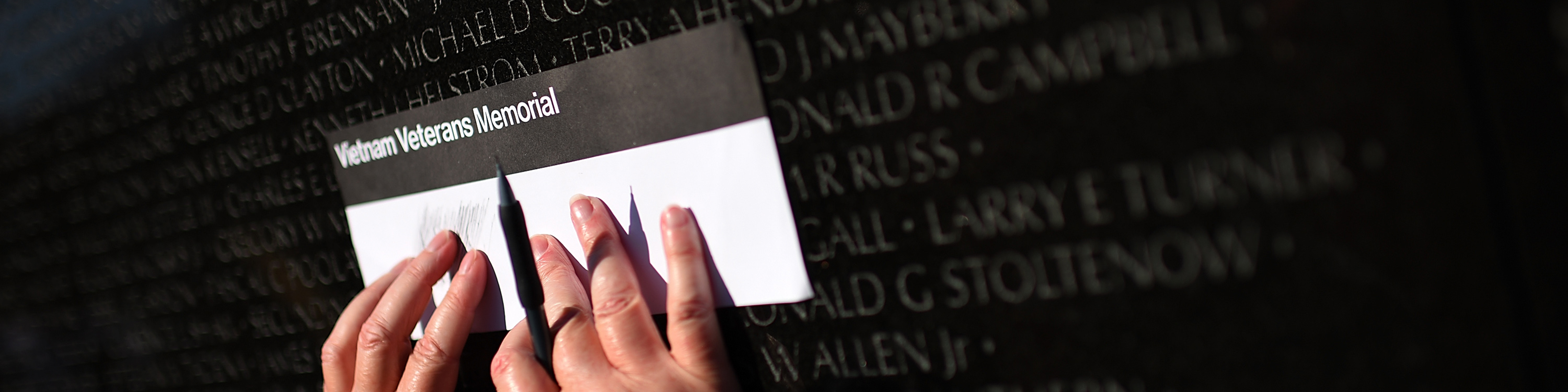 two hands hold a piece of paper up to the vietnam veterans memorial to make an etching of a name on the wall