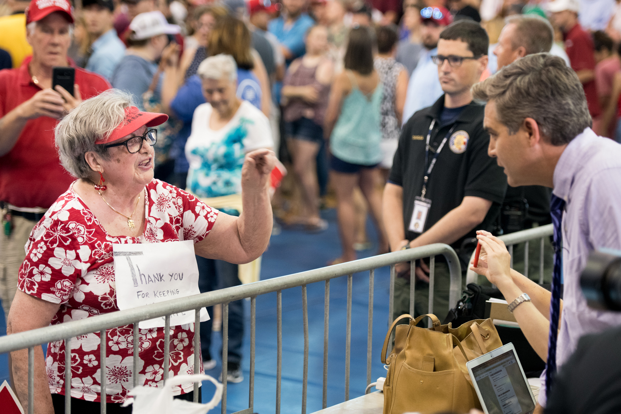 An elderly woman in a make America great again hat shouts and points at Jim Acosta, who is standing behind a metal barricade