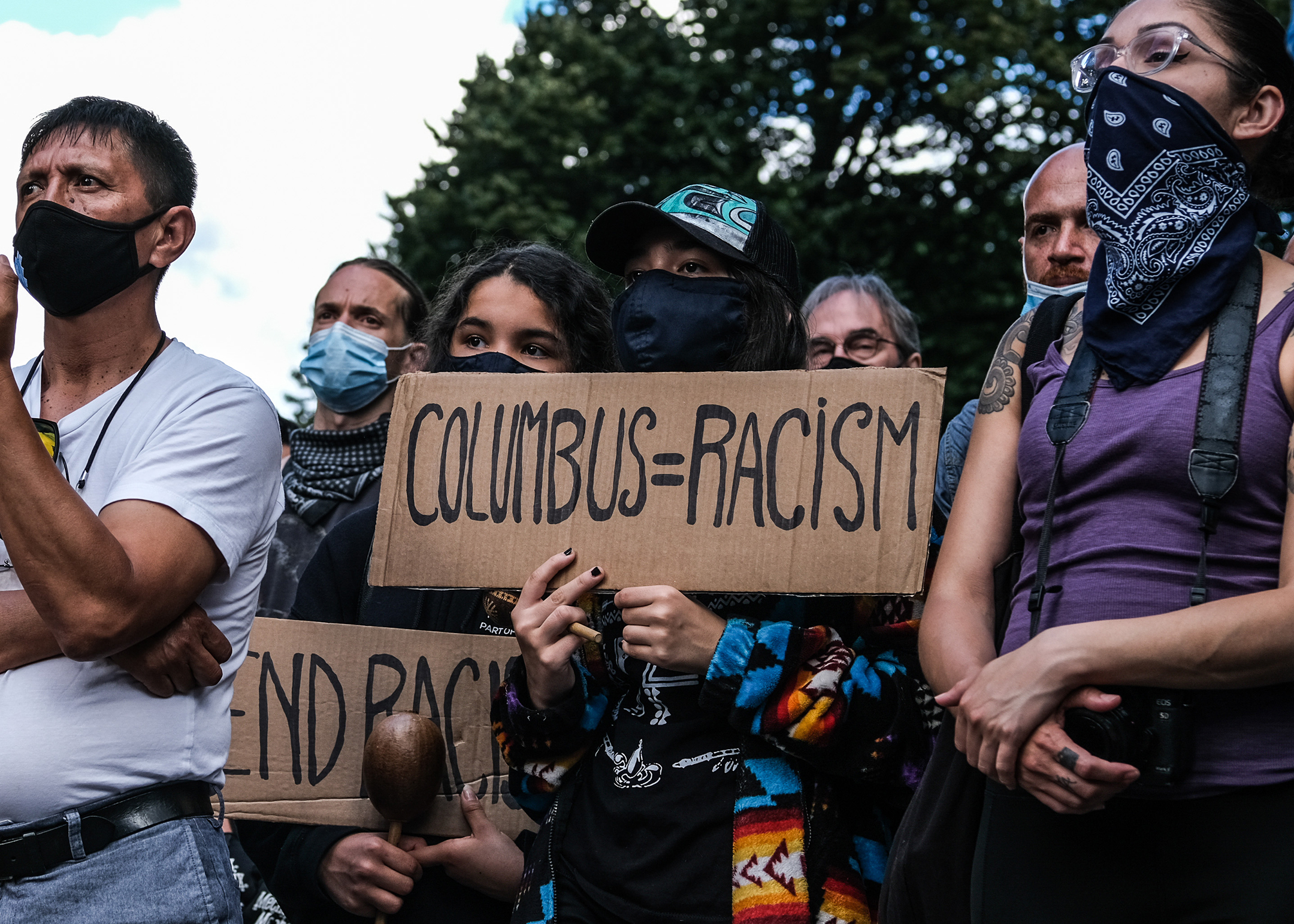A group of protesters wearing facemasks hold handmade signs that read columbus=racist