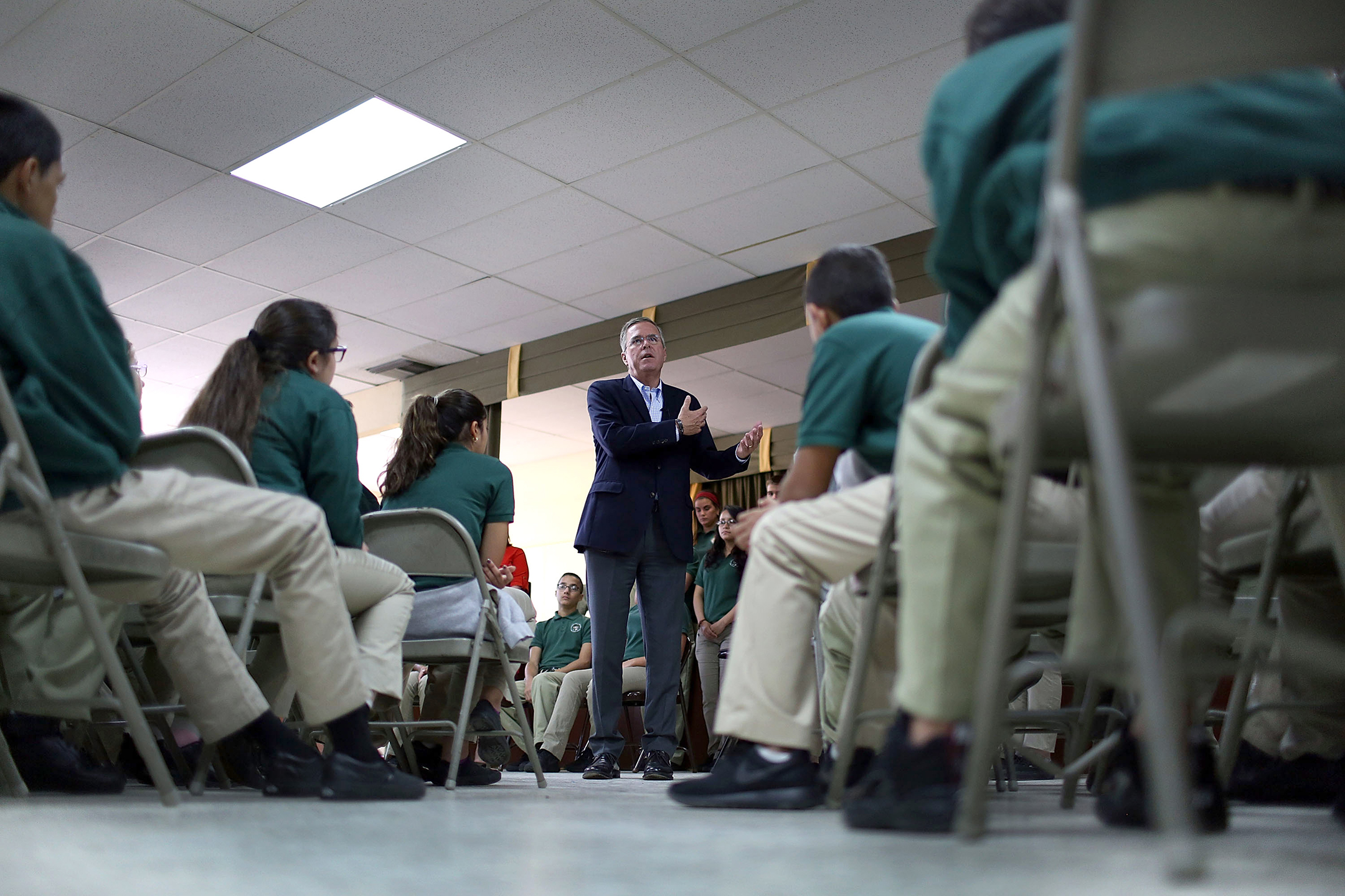 jeb bush, standing in a suit at the center of the photograph, talks with a group of students who are seated in front of him