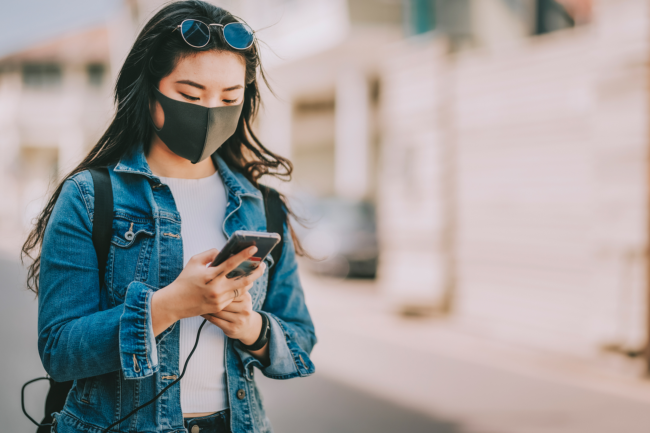 Photo of a young Asian woman wearing a face mask while standing on the street and looking at her phone