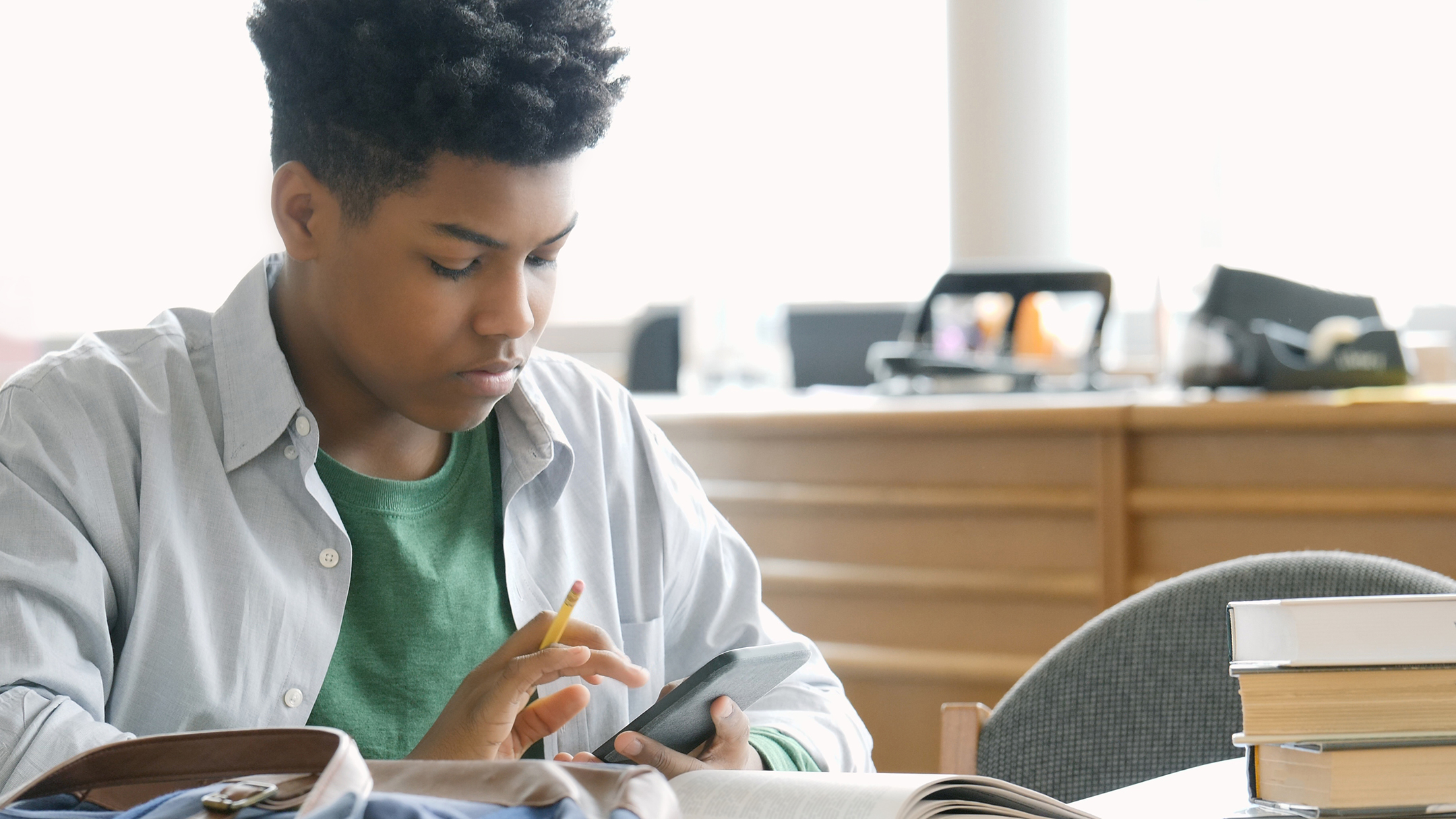 A young Black man looks at his phone wile sitting at a desk with books and homework