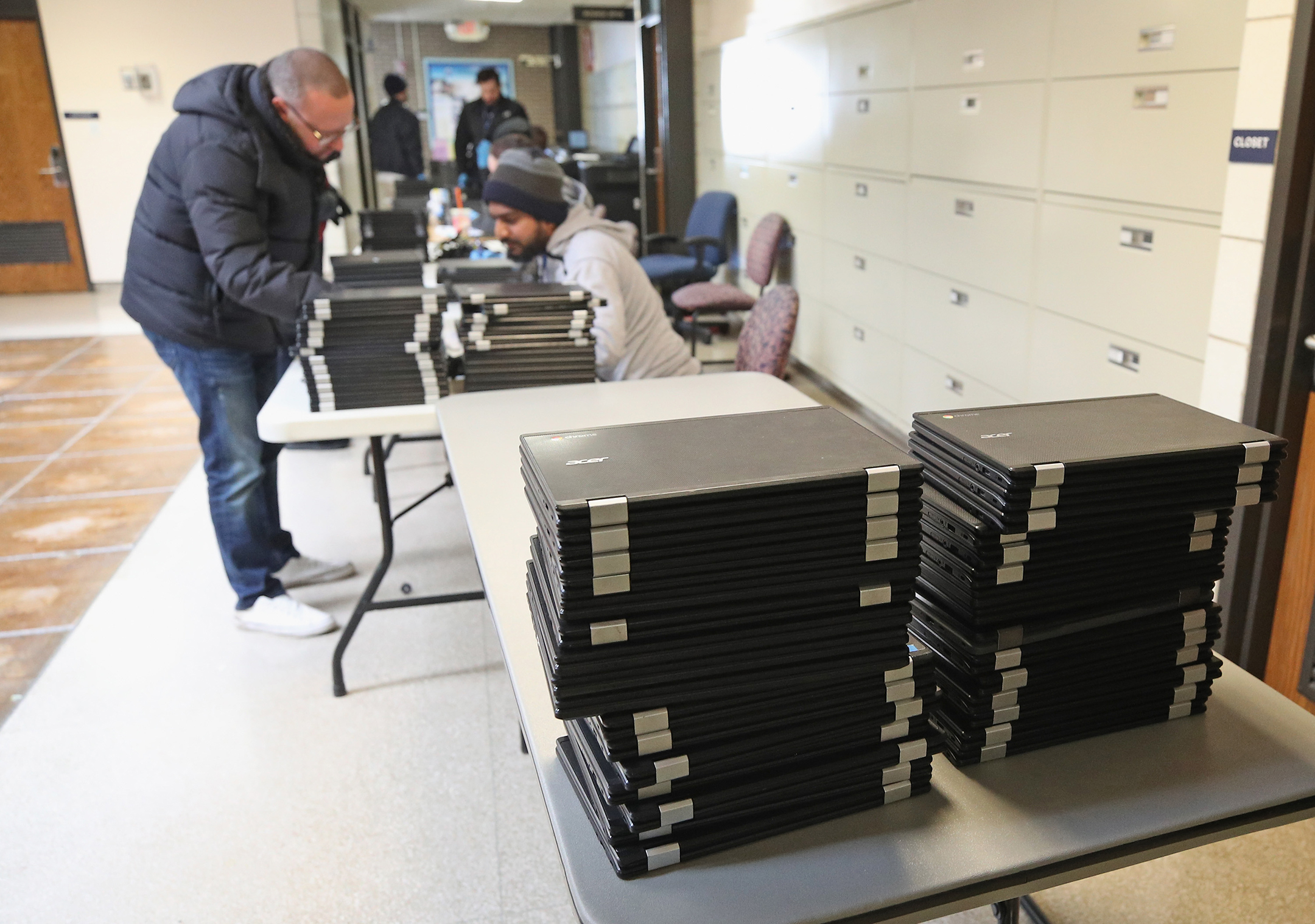 Photograph of a stack of laptop computers in the foreground, with an educator helping a parent check out a computer in the background