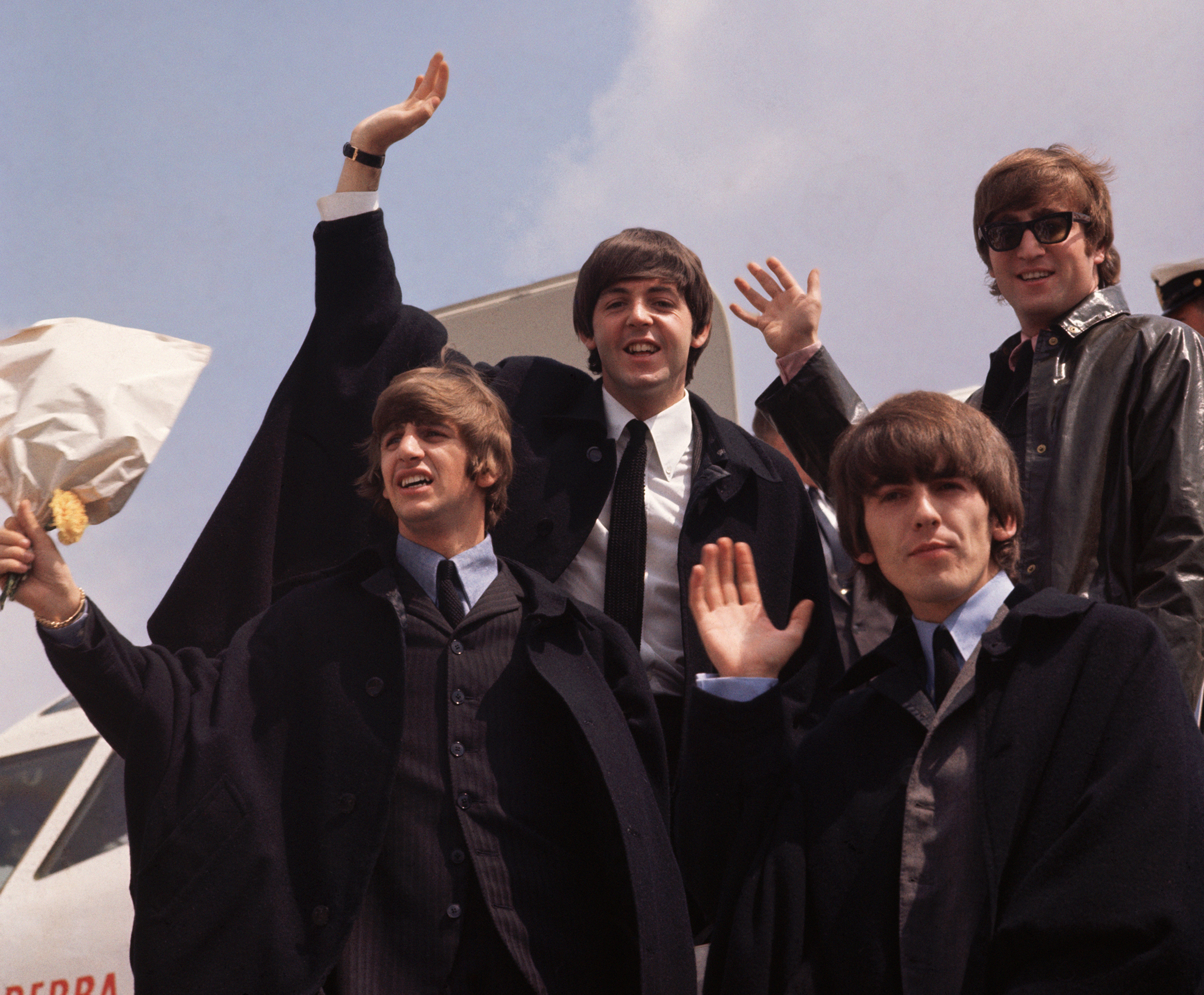The Beatles smiling and waving from the top of a set of steps attached to an airplane