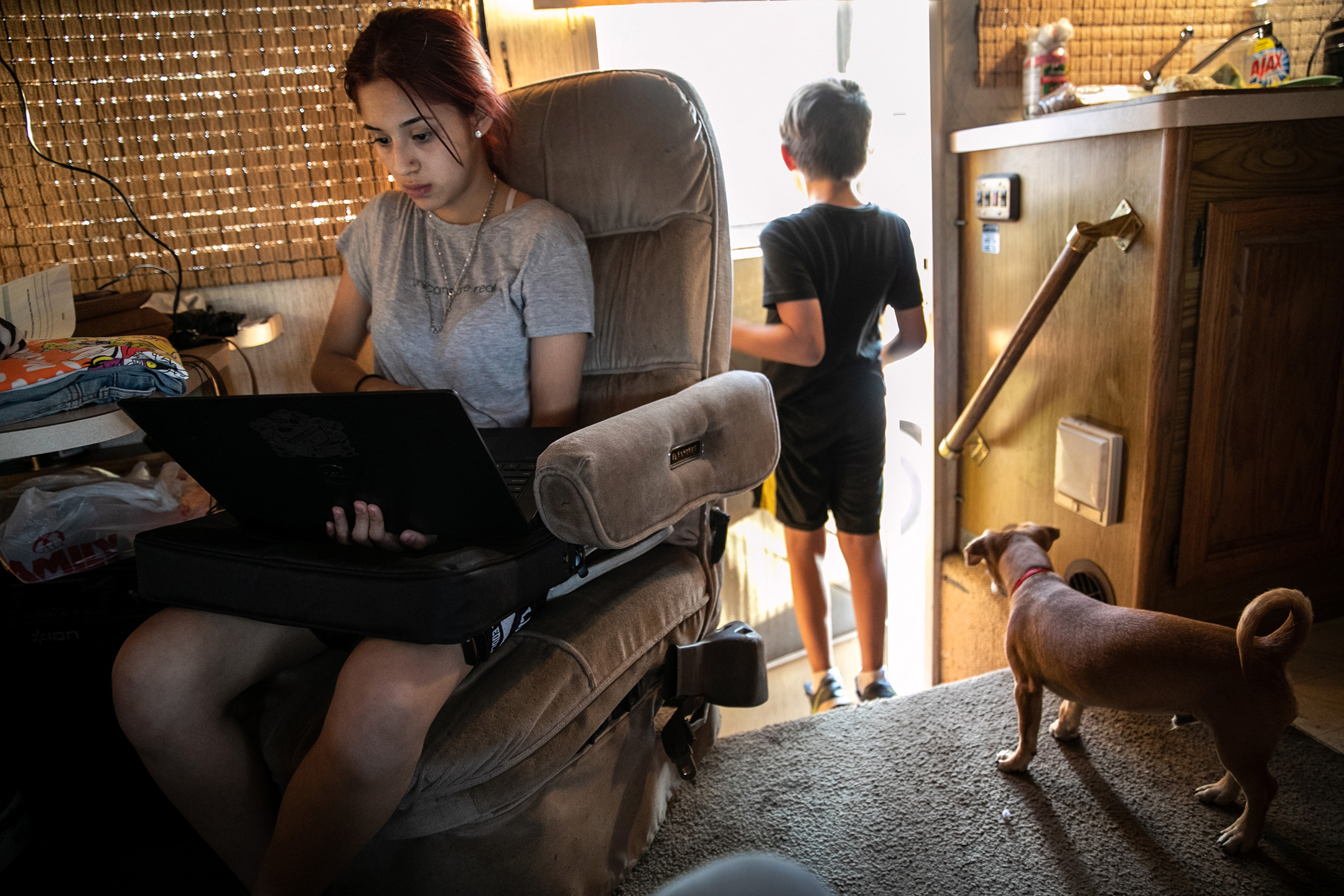 A young woman sits in a recliner with a laptop computer near the open door inside a cluttered RV