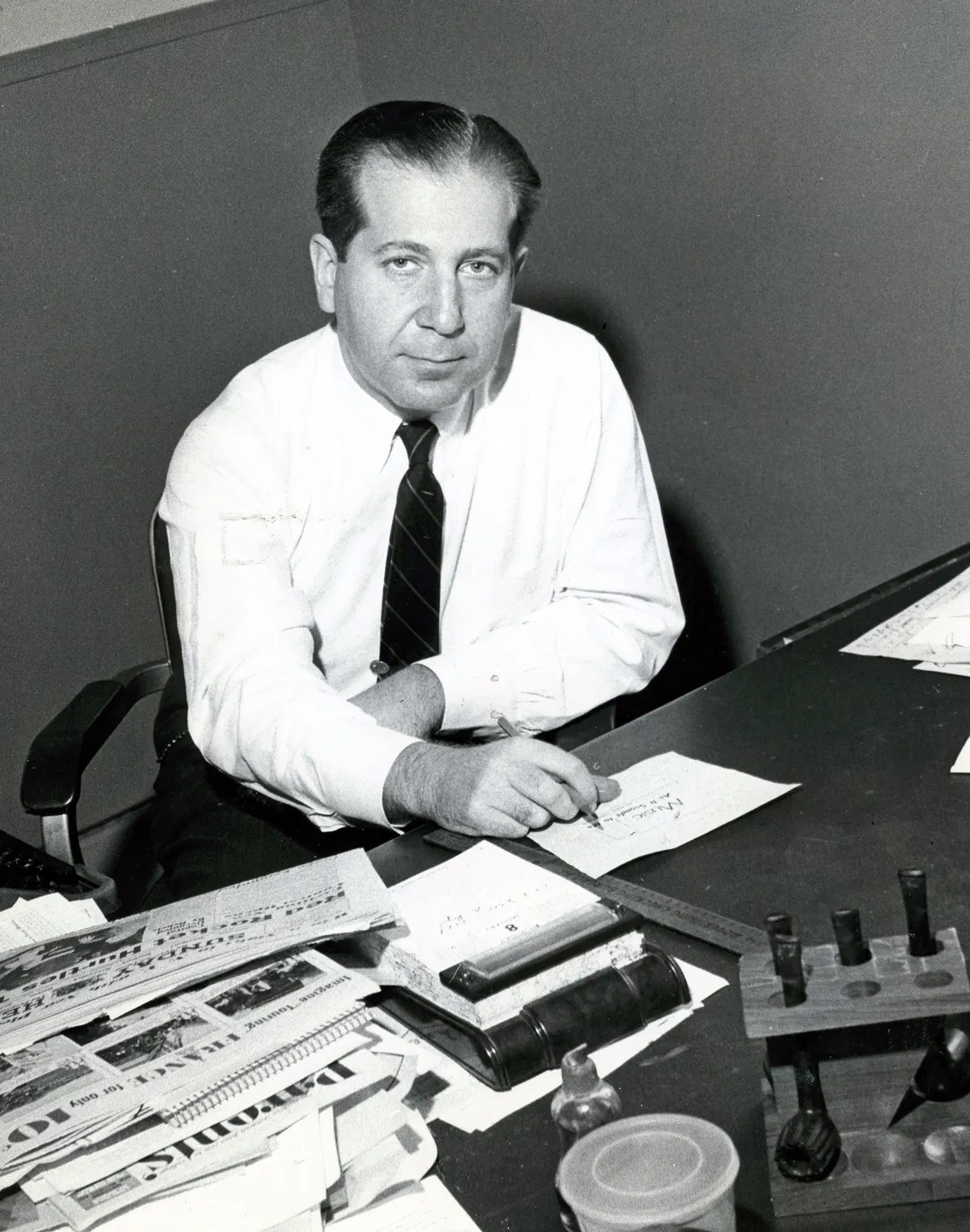 Black and white photo of journalist Fred M. Hechinger sitting at his desk, which has a pile of newspapers on it