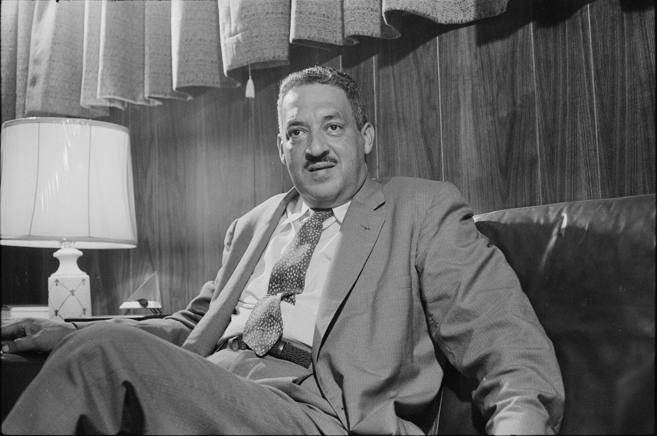 Thurgood Marshall in a suit sits on a leather couch in a wood-paneled room