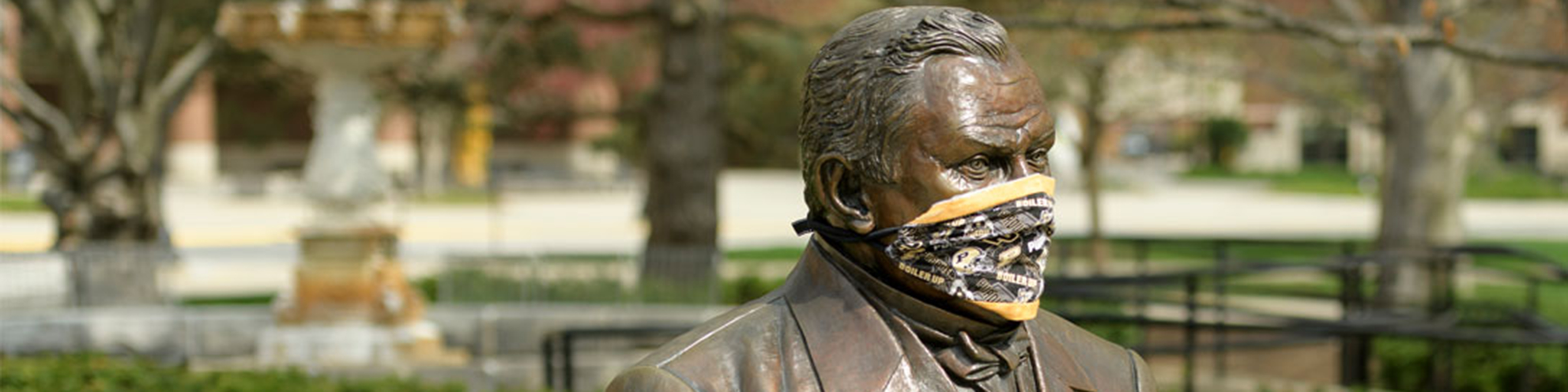 Photograph of a bronze statue on the campus of Perdue university with a Perdue facemask around his face