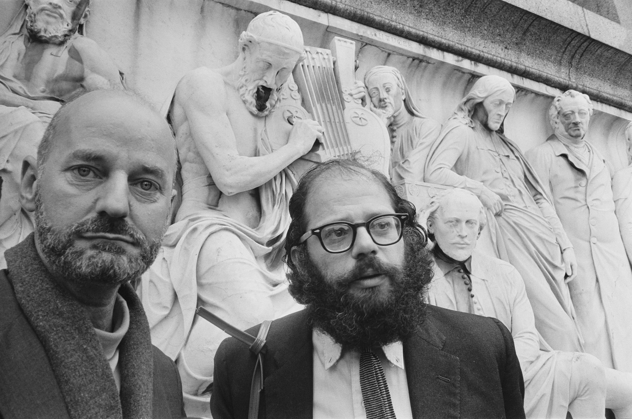 Black and white photo of Lawrence Ferlinghetti and Allen Ginsberg standing in front of a frieze, with Ferlinghetti looking at the camera and Ginsberg looking slightly to his left