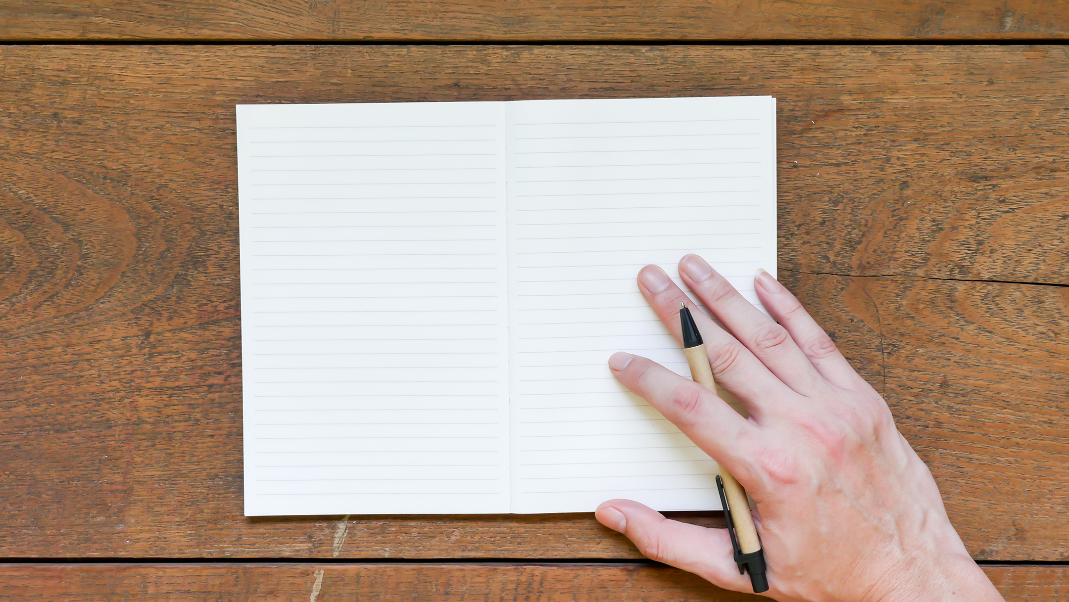 open blank journal on a wooden table, with a hand holding a pencil on the right side of the journal
