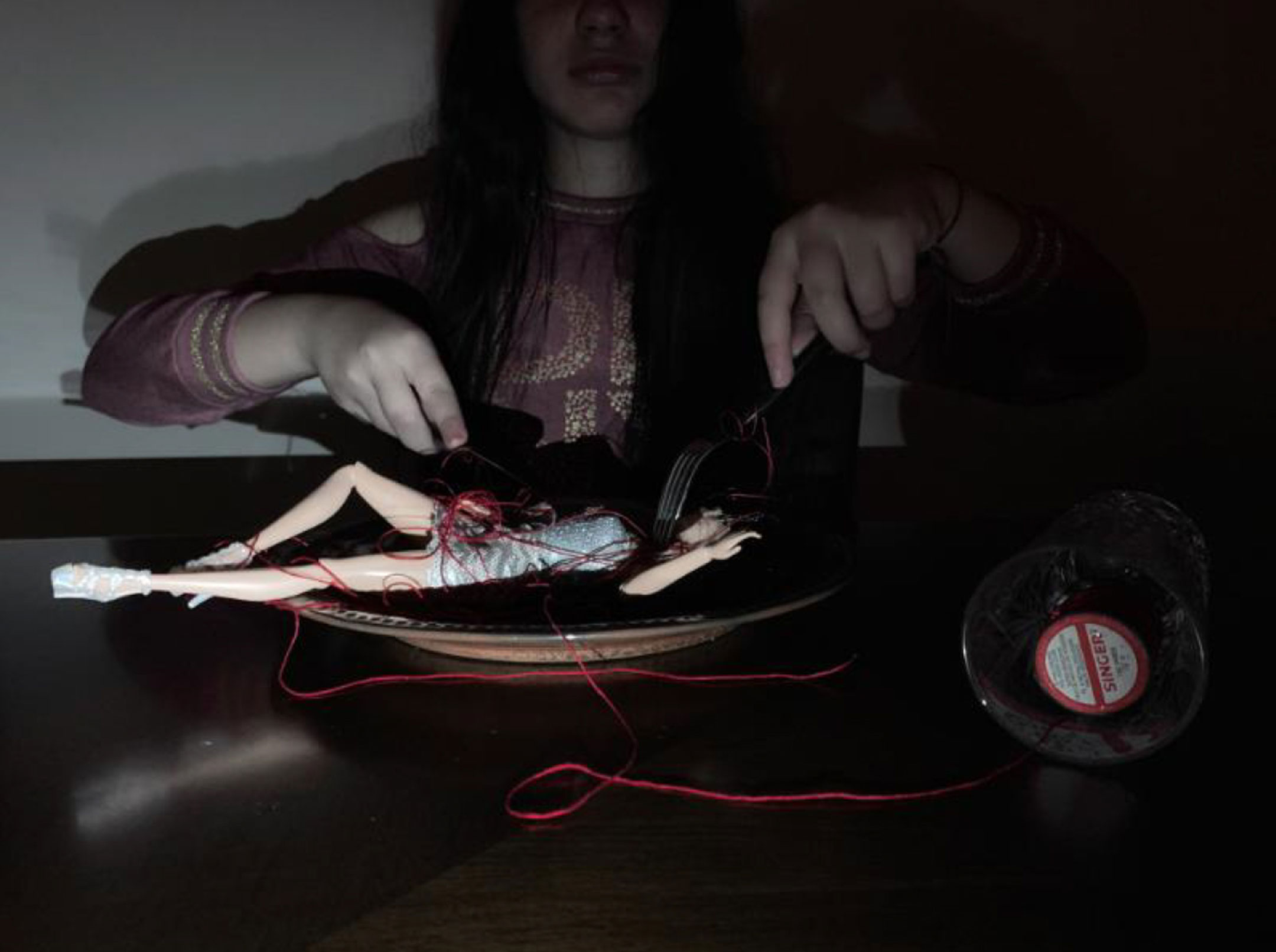 Dark photograph of a girl holding a fork and knife as she pretends to eat a Barbie doll
