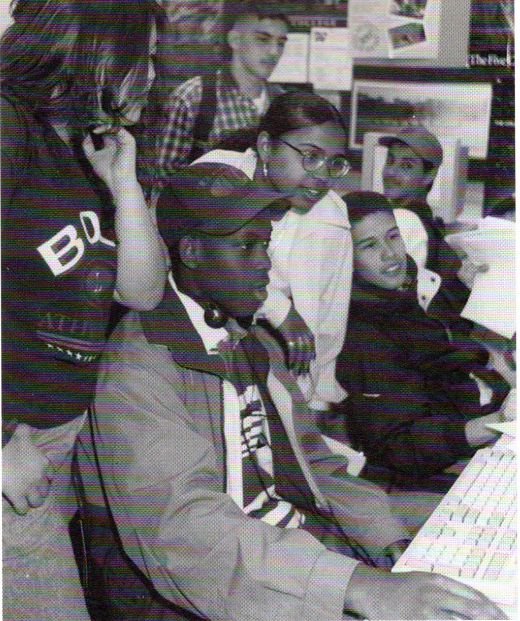 Black and white photo of a group of black high school students, male and female, gathered around a computer, which a young black man sitting in front of the machine