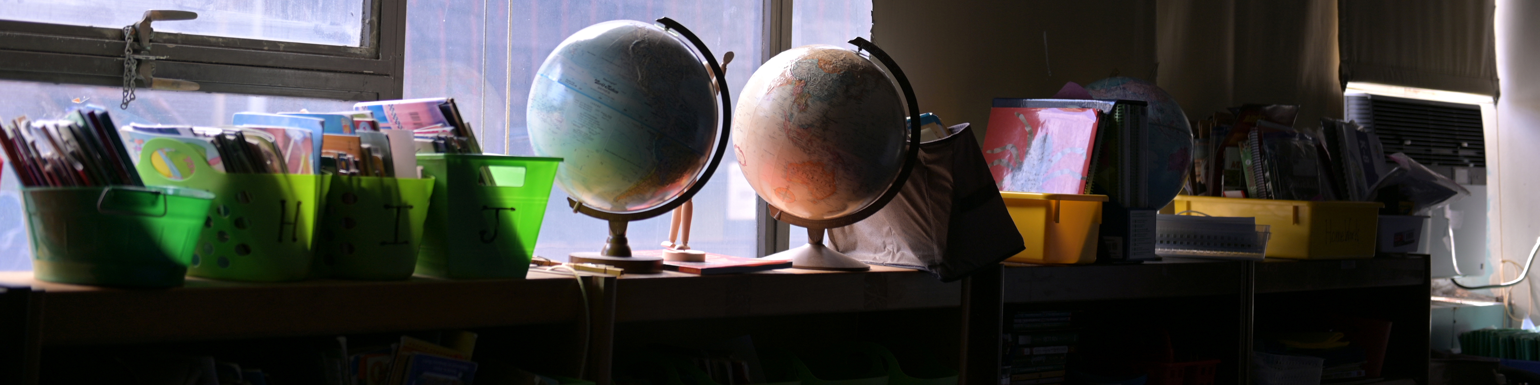A classroom is empty with the lights off with books, globes, and other supplies seen in shadow against a window
