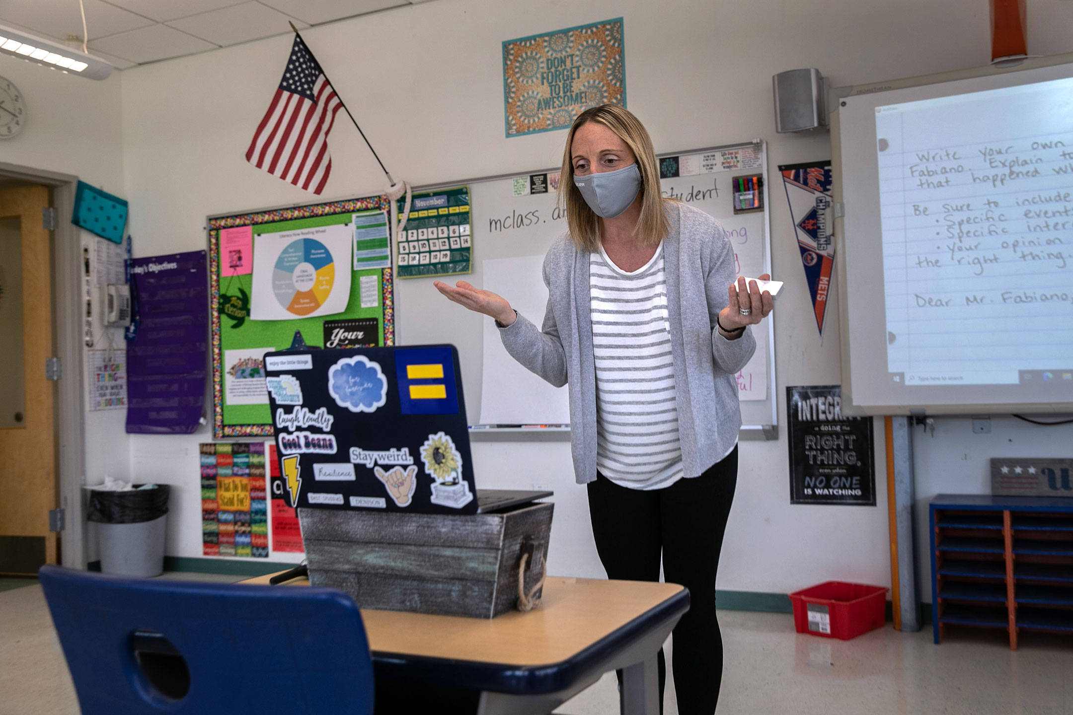 White teacher wearing a mask stands in front of her laptop and speaks to her remote class