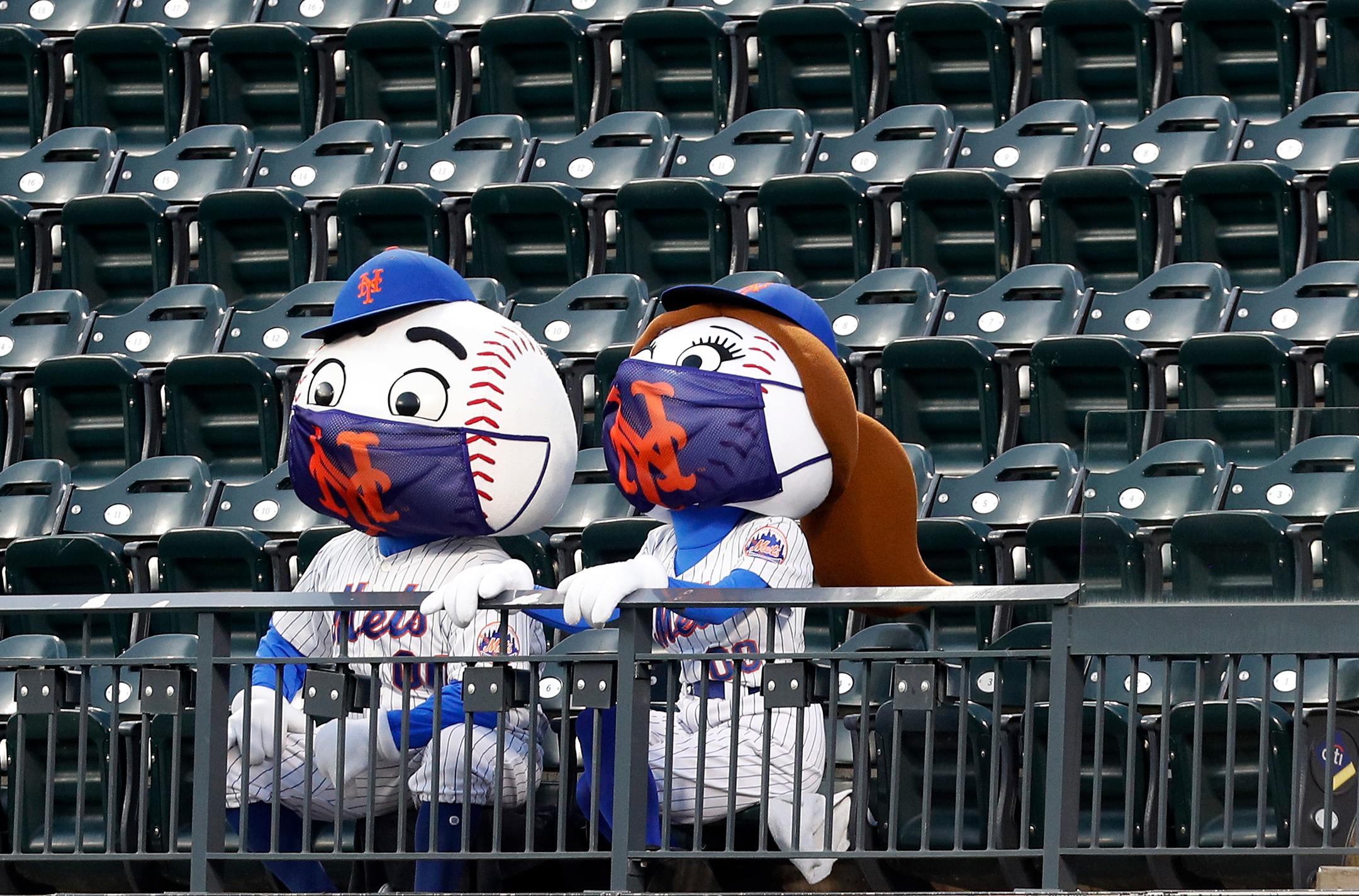 Mr. and Mrs. Met sit next to each other while wearing masks in an empty stadium