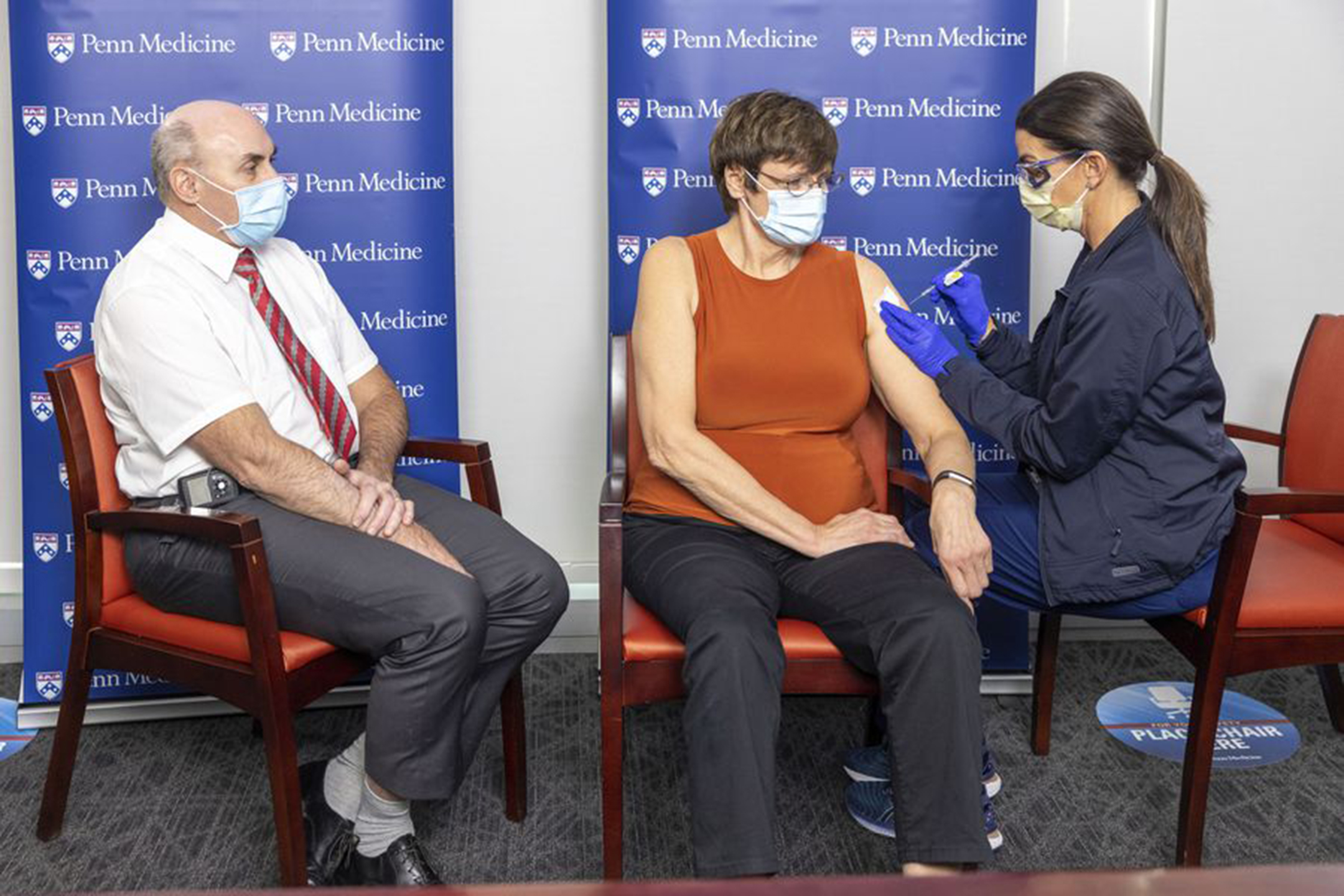 Three people sitting, all wearing masks. A man on the left, a woman in the middle getting a shot, and a nurse of the right administering the shot.