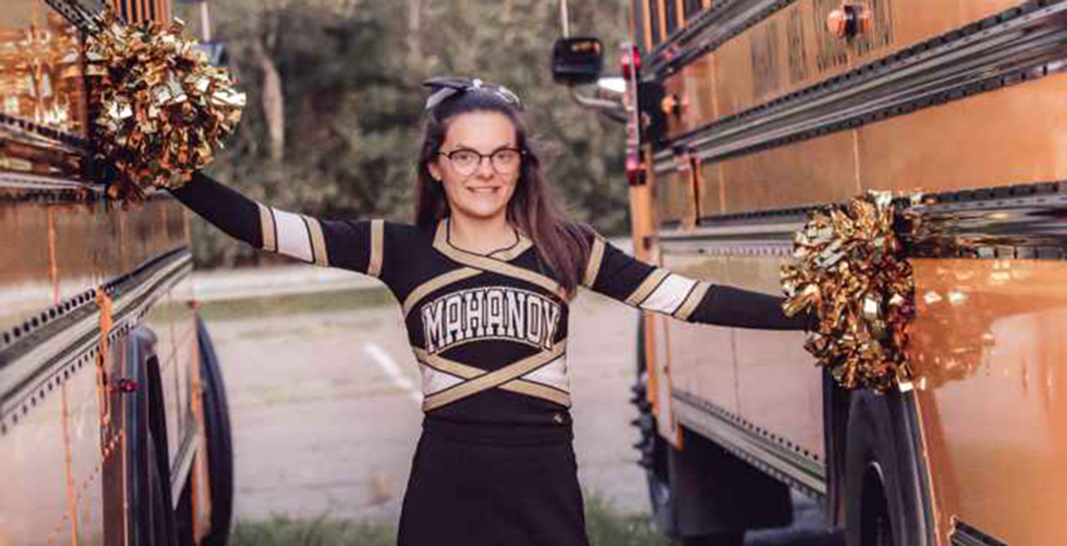 Young woman in a cheerleader outfit smiling and holding pom poms while standing between two school busses