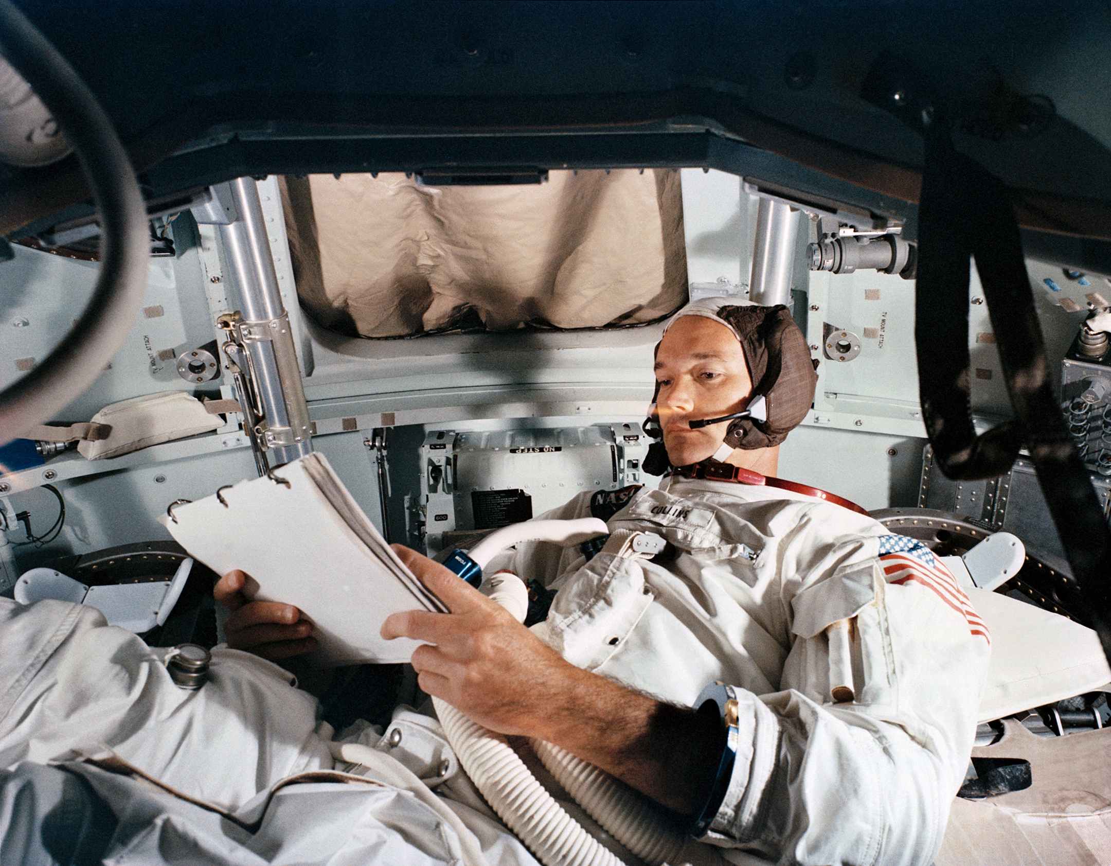 Astronaut Michael Collins, in his space suit, looking over documents in a space capsule
