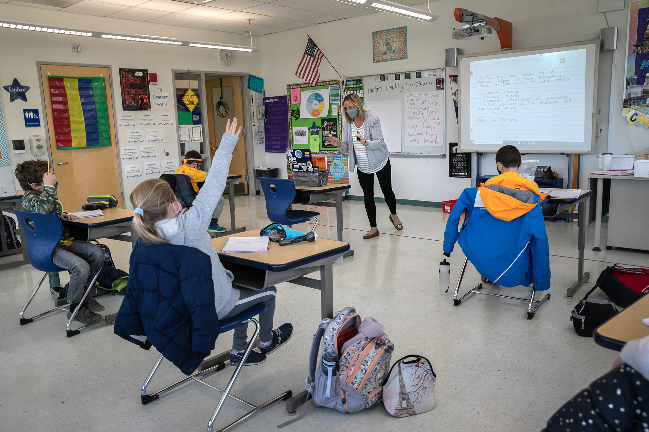 A teacher wearing a mask stands in front of a laptop in a classroom with four students, one with their hand raised