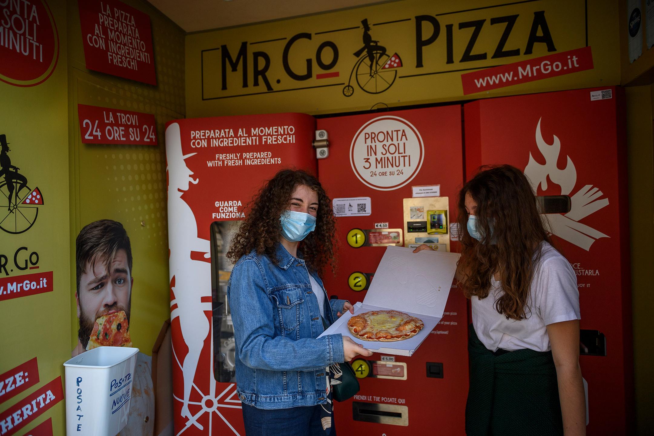 Two young women, both wearing face masks, hold a pizza in front of a red pizza vending machine